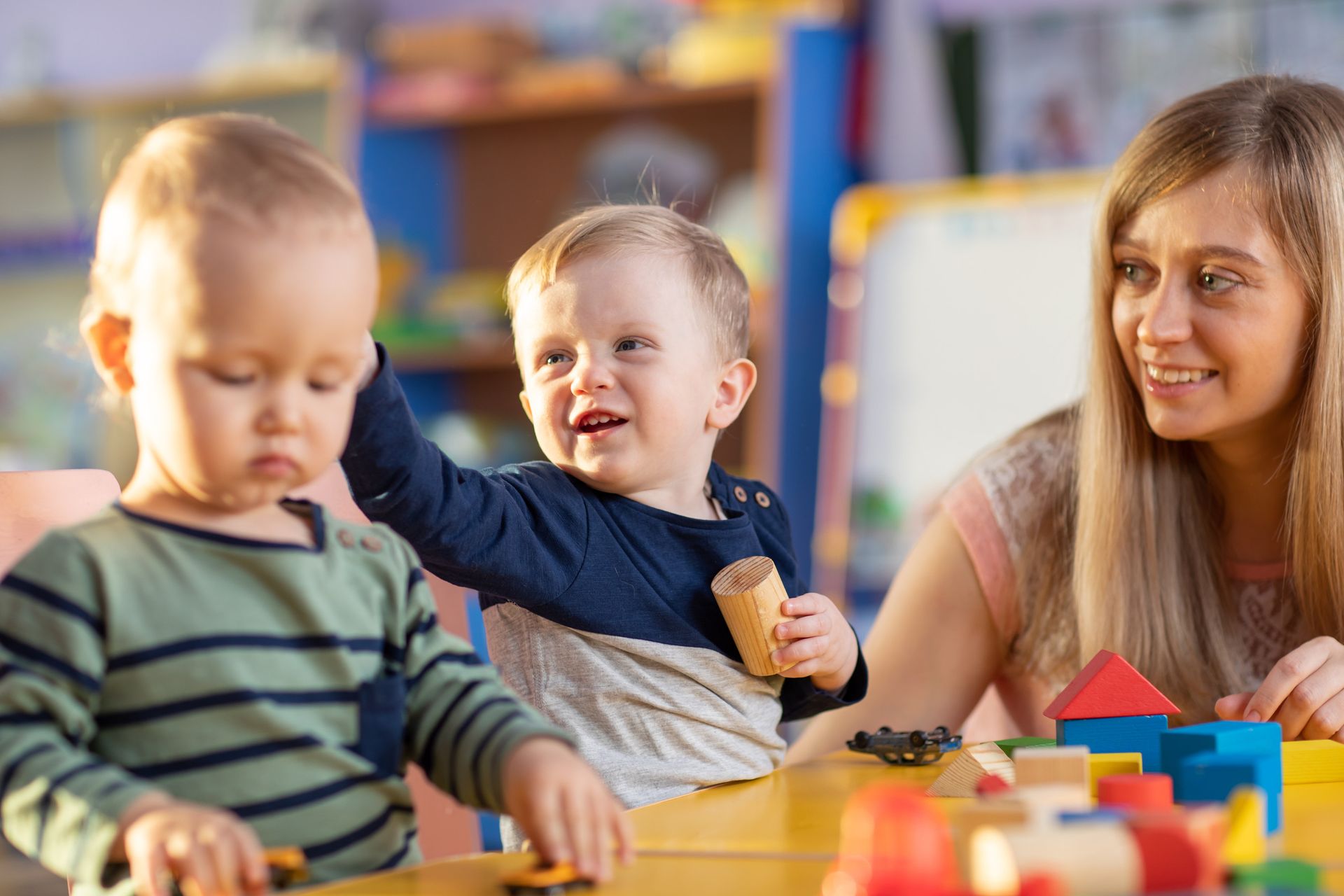 A woman smiles at two toddlers playing with toys and building blocks at a table.