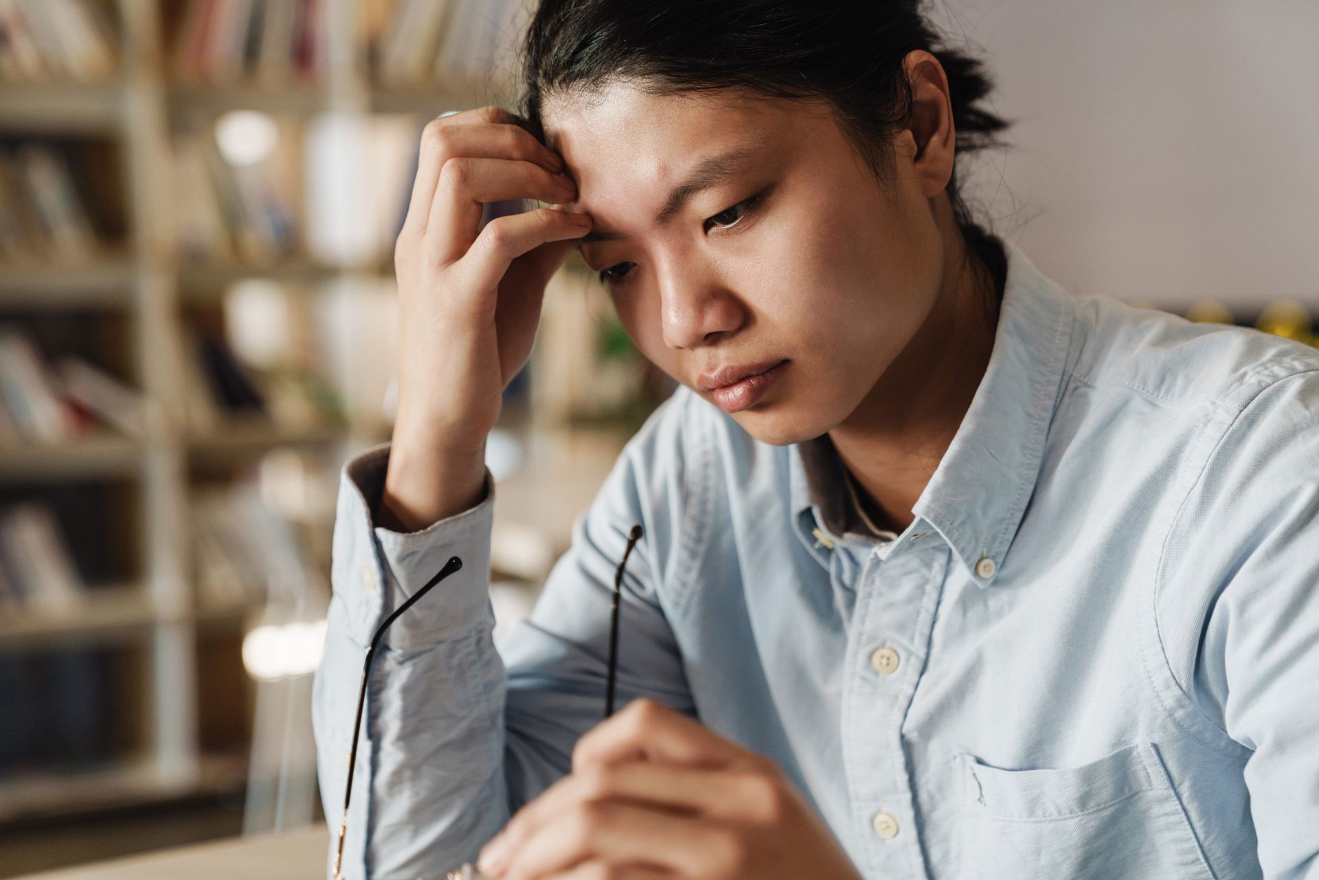 Asian person in light blue shirt, holding glasses, looking down with a concerned expression in a library.