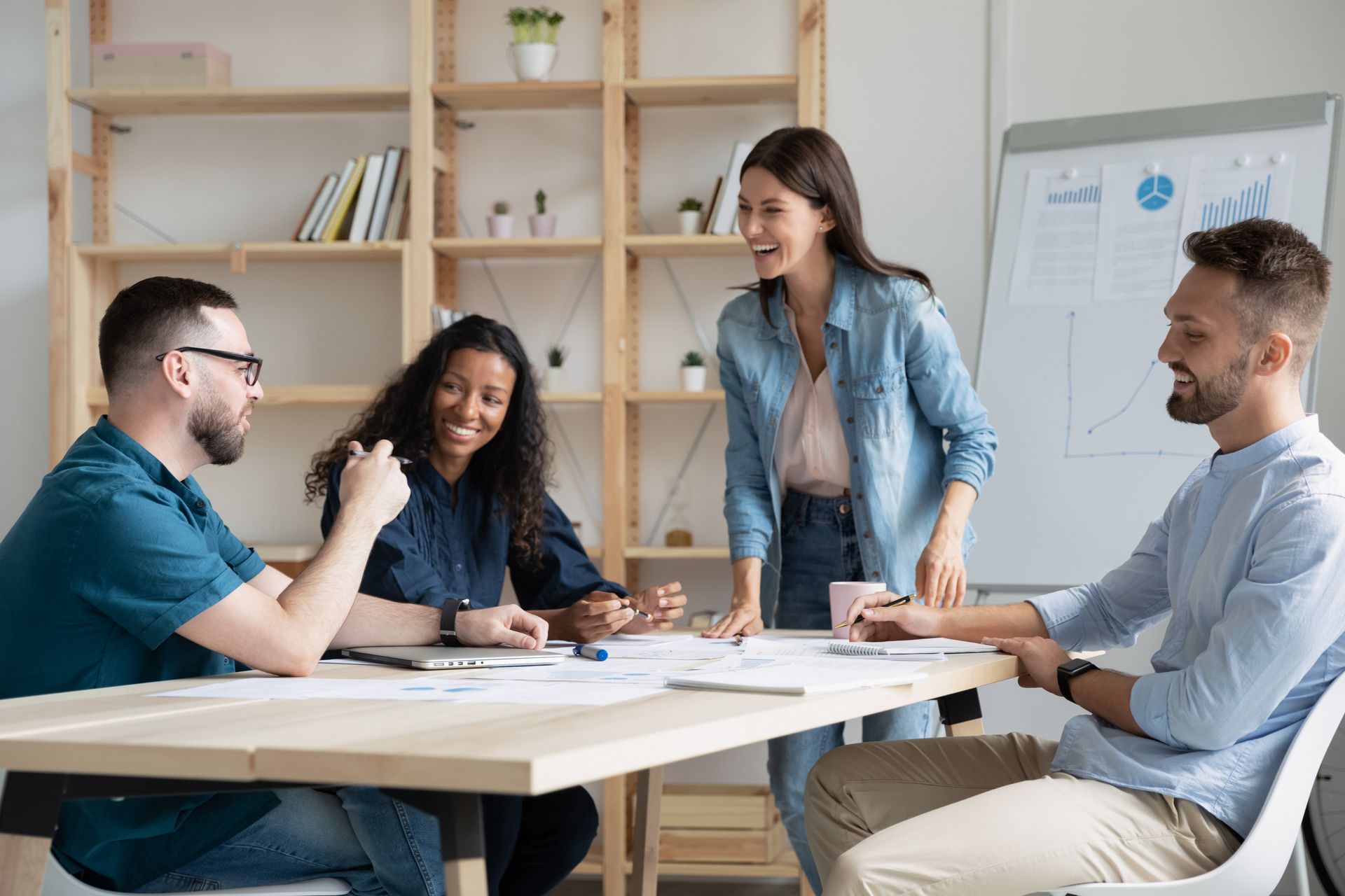 Four diverse colleagues in a bright office, laughing and collaborating around a table with documents.