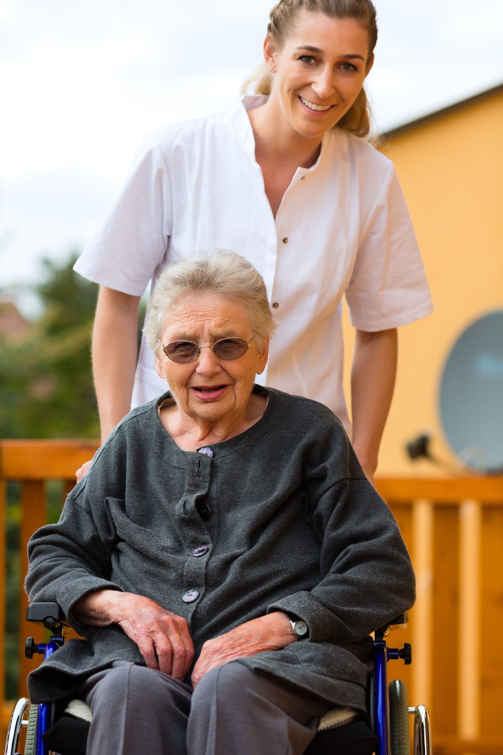 Smiling caregiver pushes elderly woman in wheelchair on outdoor deck.