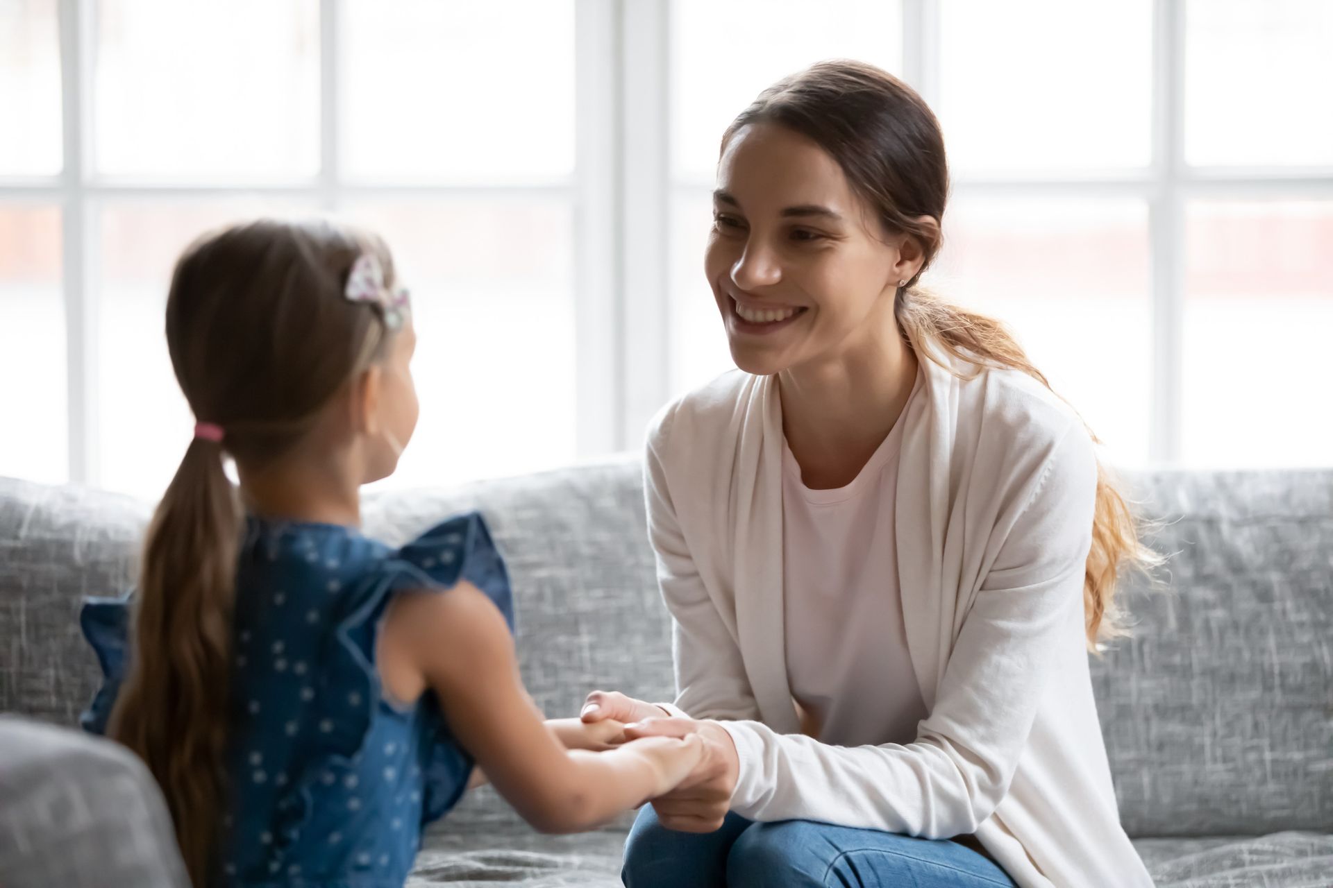 Woman smiling, holding hands with a young girl indoors on a couch, conversing.