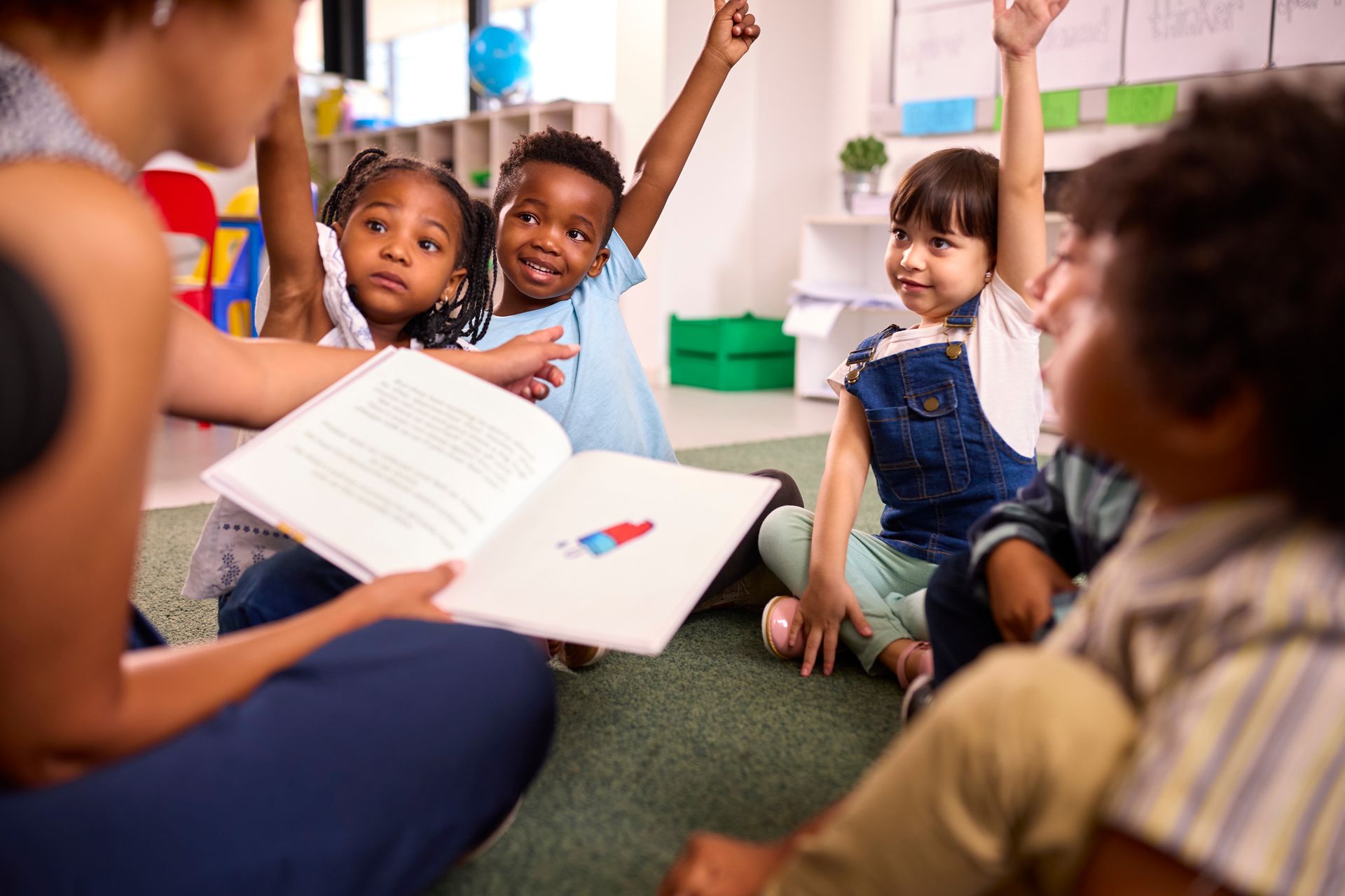 Teacher reading aloud to a group of diverse children with raised hands, in a classroom.