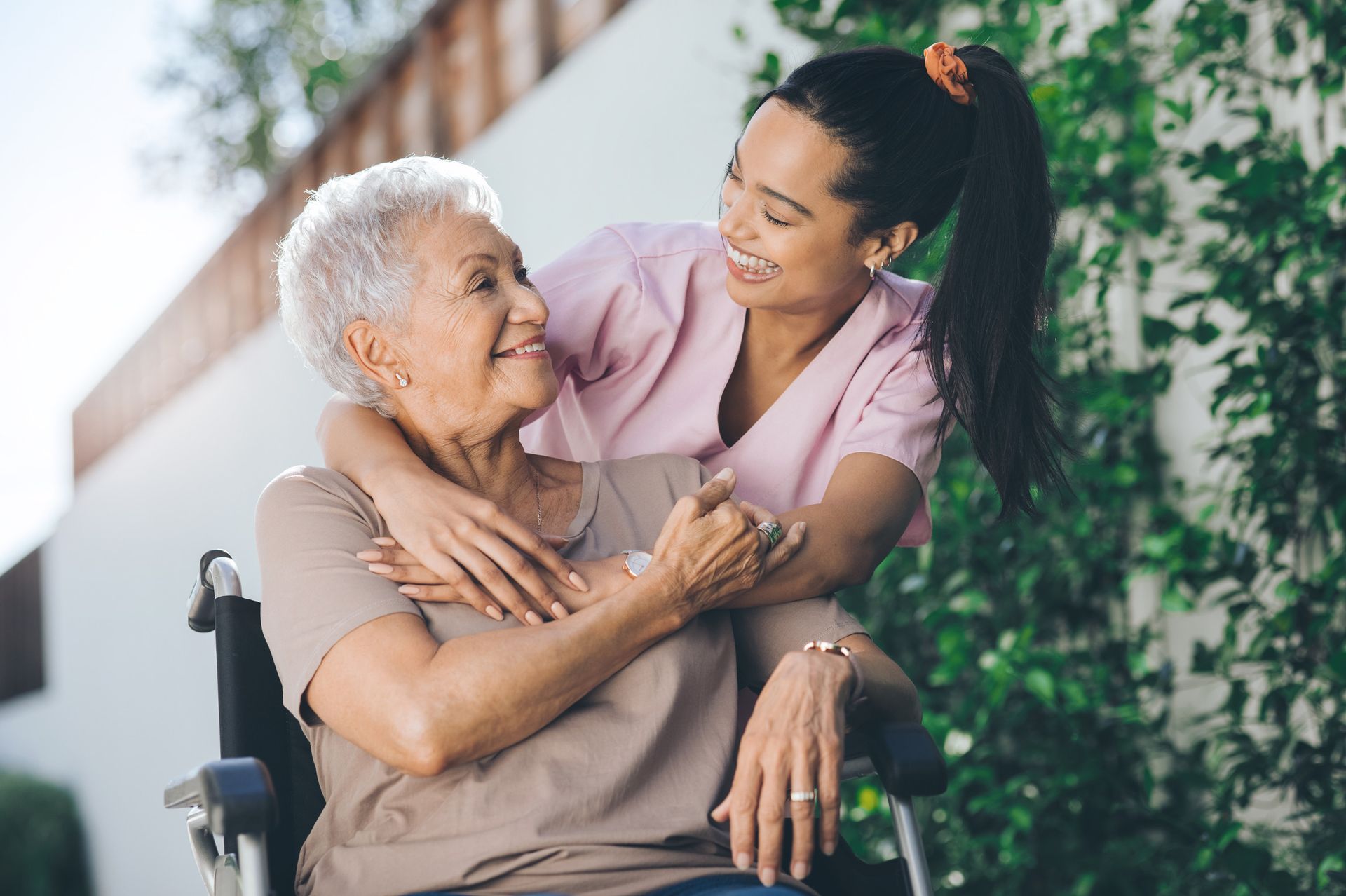 Young woman hugs an elderly woman in a wheelchair outdoors, smiling at each other.