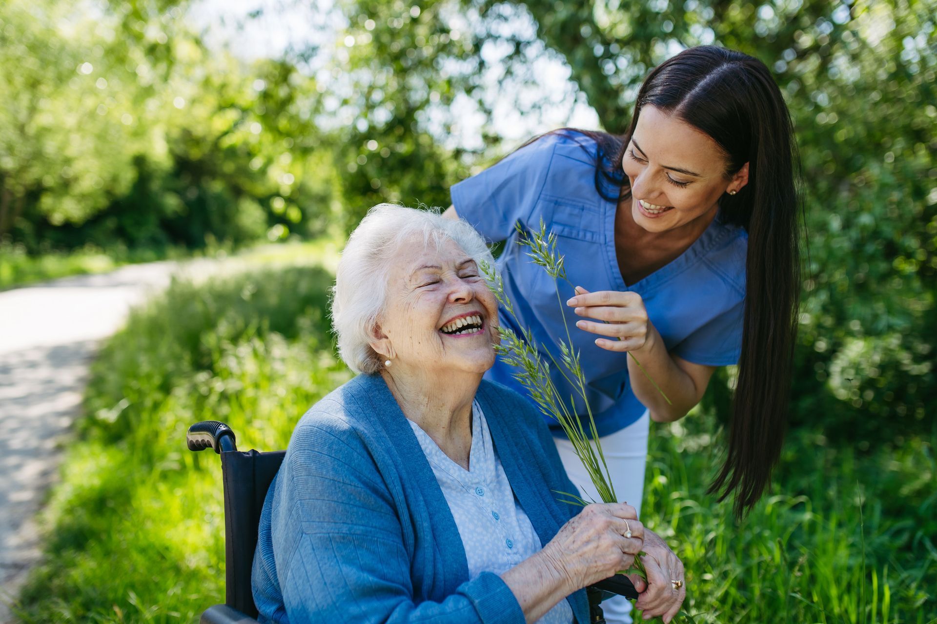 Woman in blue scrubs smiles at a laughing elderly woman in a wheelchair, outdoors with greenery.