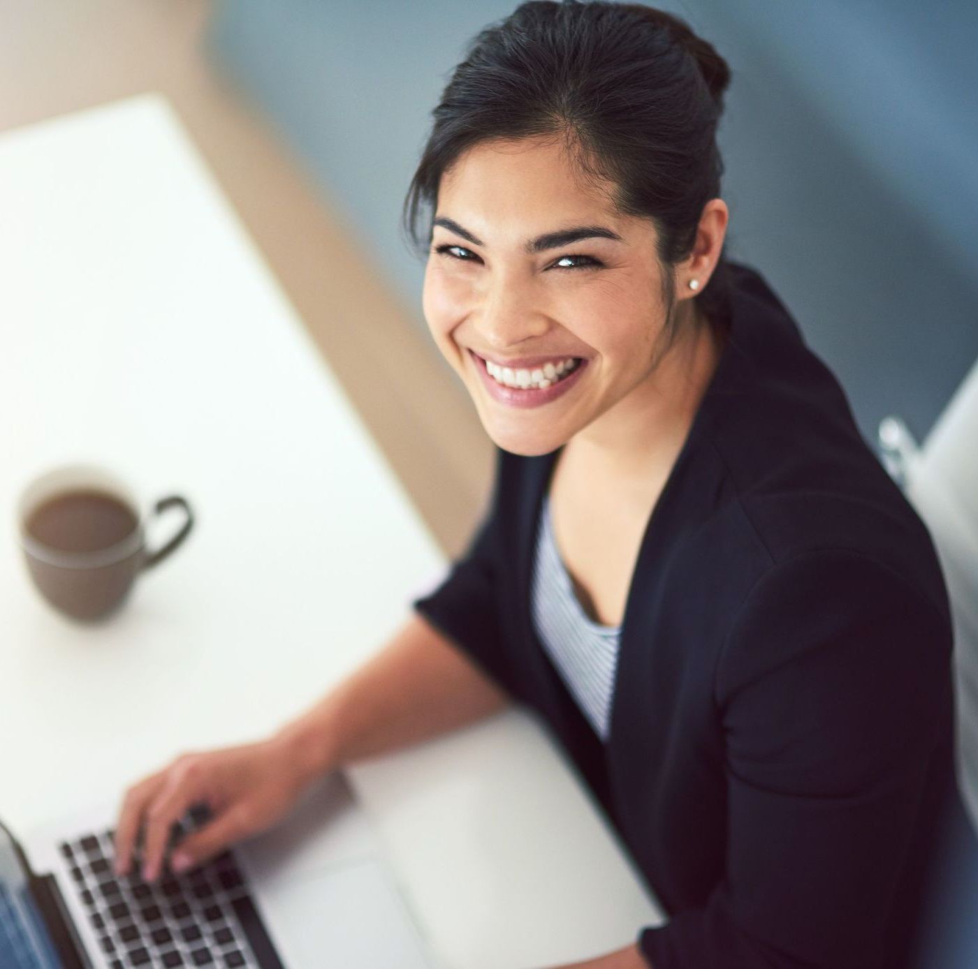 Woman smiles at camera while working on a laptop at a white table.