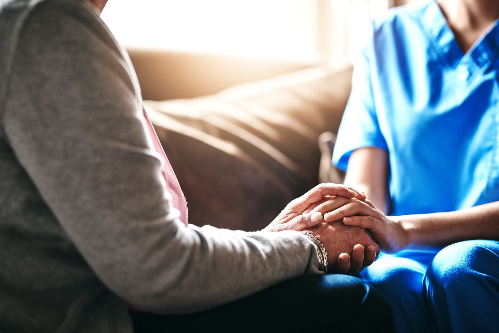 A healthcare worker in blue scrubs holds the hands of an elderly person for comfort.