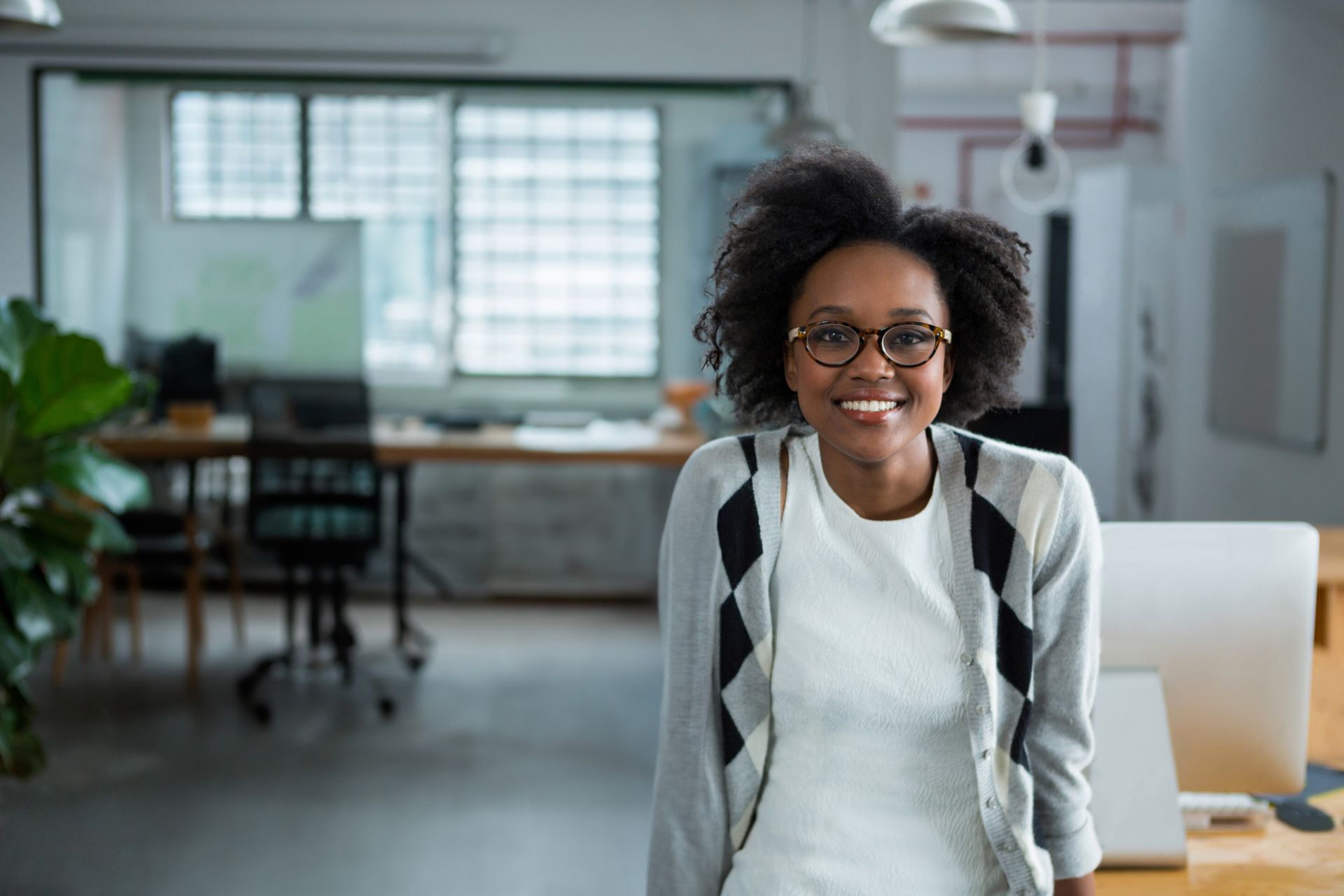 Woman with glasses smiling in an office. She wears a gray cardigan and white shirt.