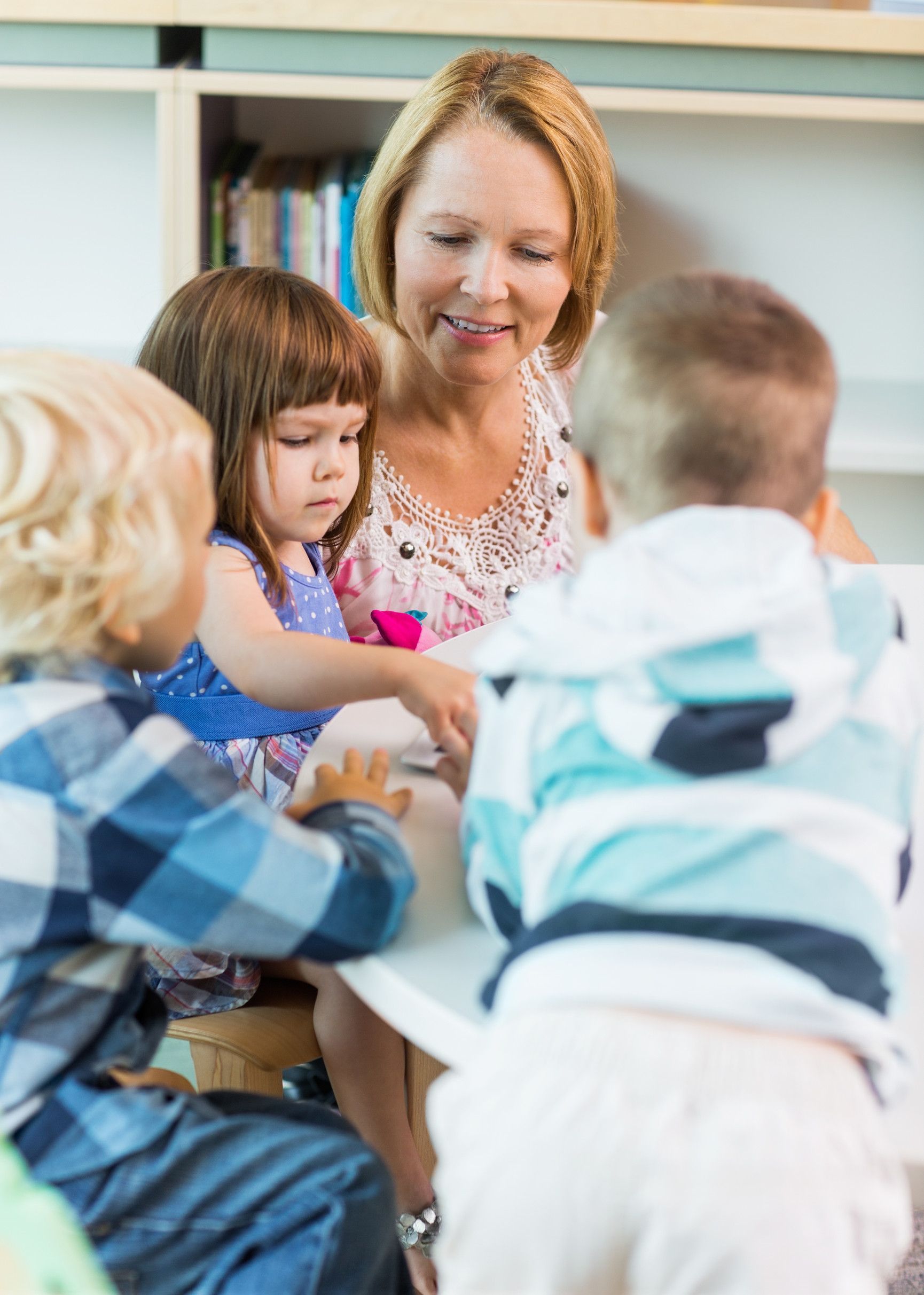 A woman with blond hair sits with three children at a table. She smiles at them, and they look at something on the table.