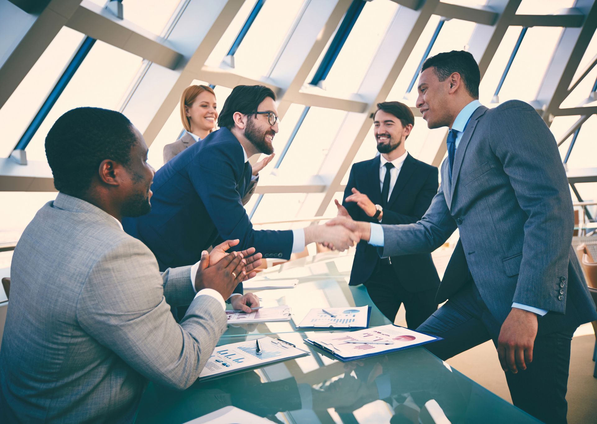 Business people in suits shaking hands in a modern office, others watching and applauding.