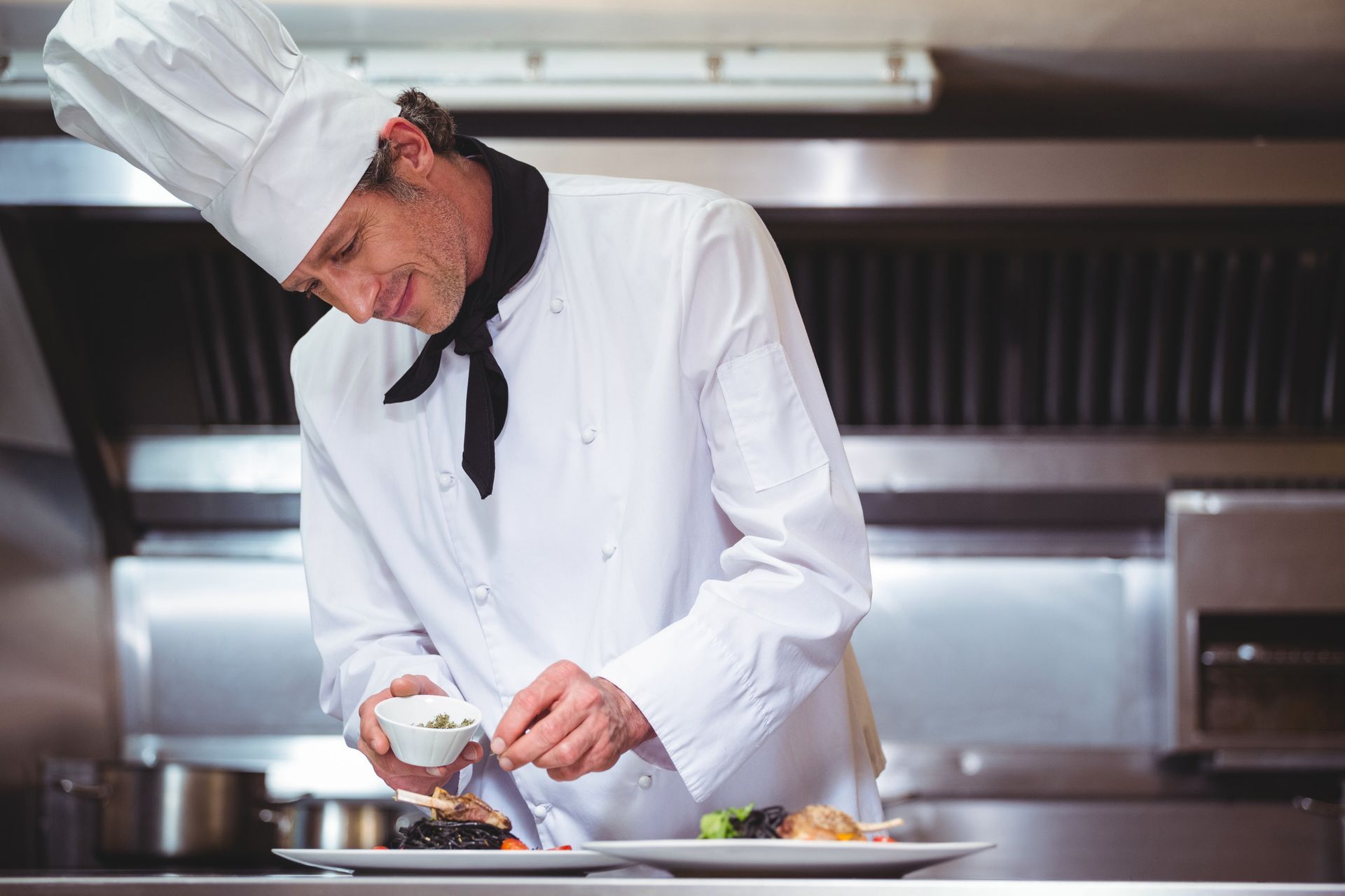 Chef in white uniform plating food in a commercial kitchen, adding garnish.