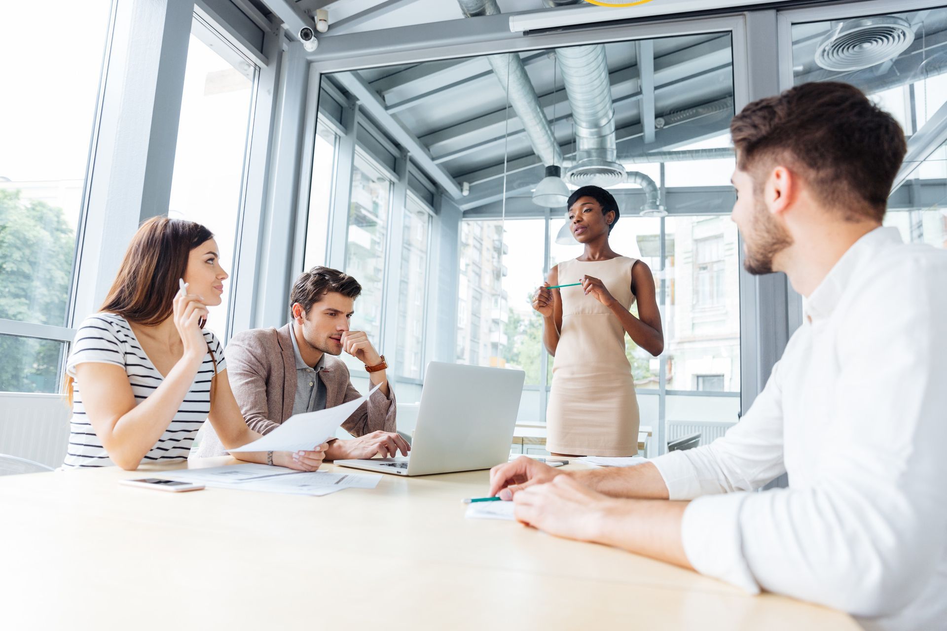 Four people in a bright room, in a meeting; one woman speaks, three others listen and look at laptop.