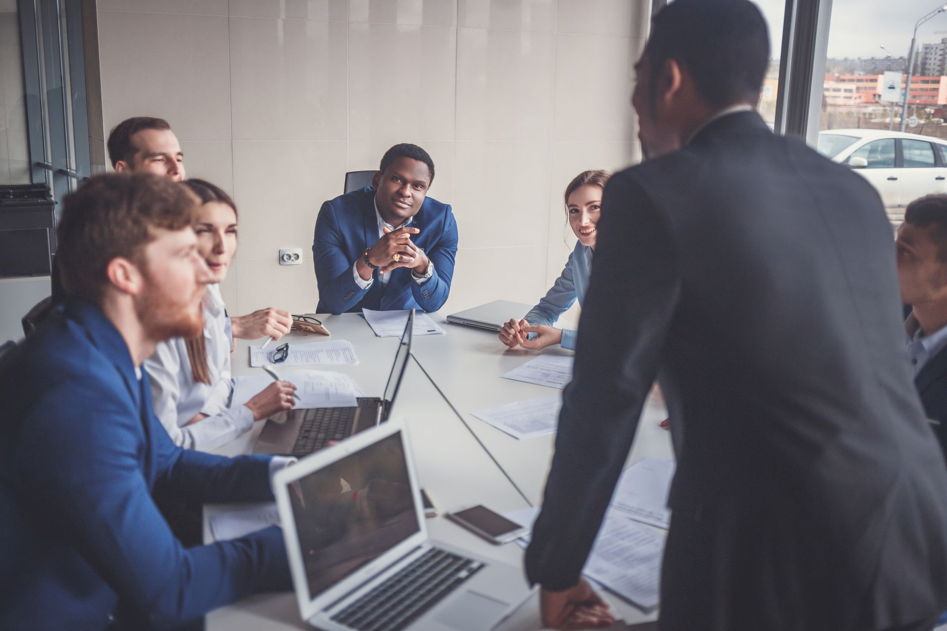 Business meeting in progress; diverse group seated around a table, listening to a man gesturing and speaking.