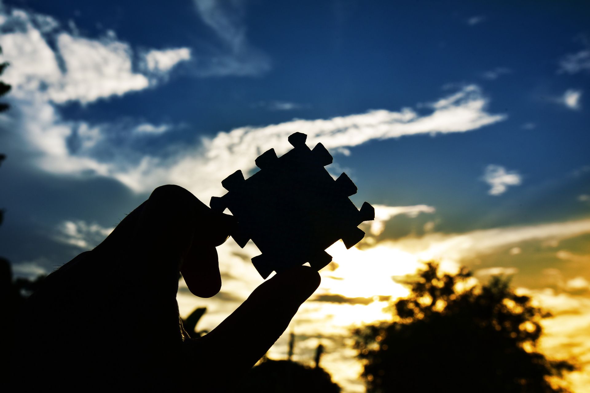 Silhouette of hand holding a puzzle piece against a colorful sunset sky.