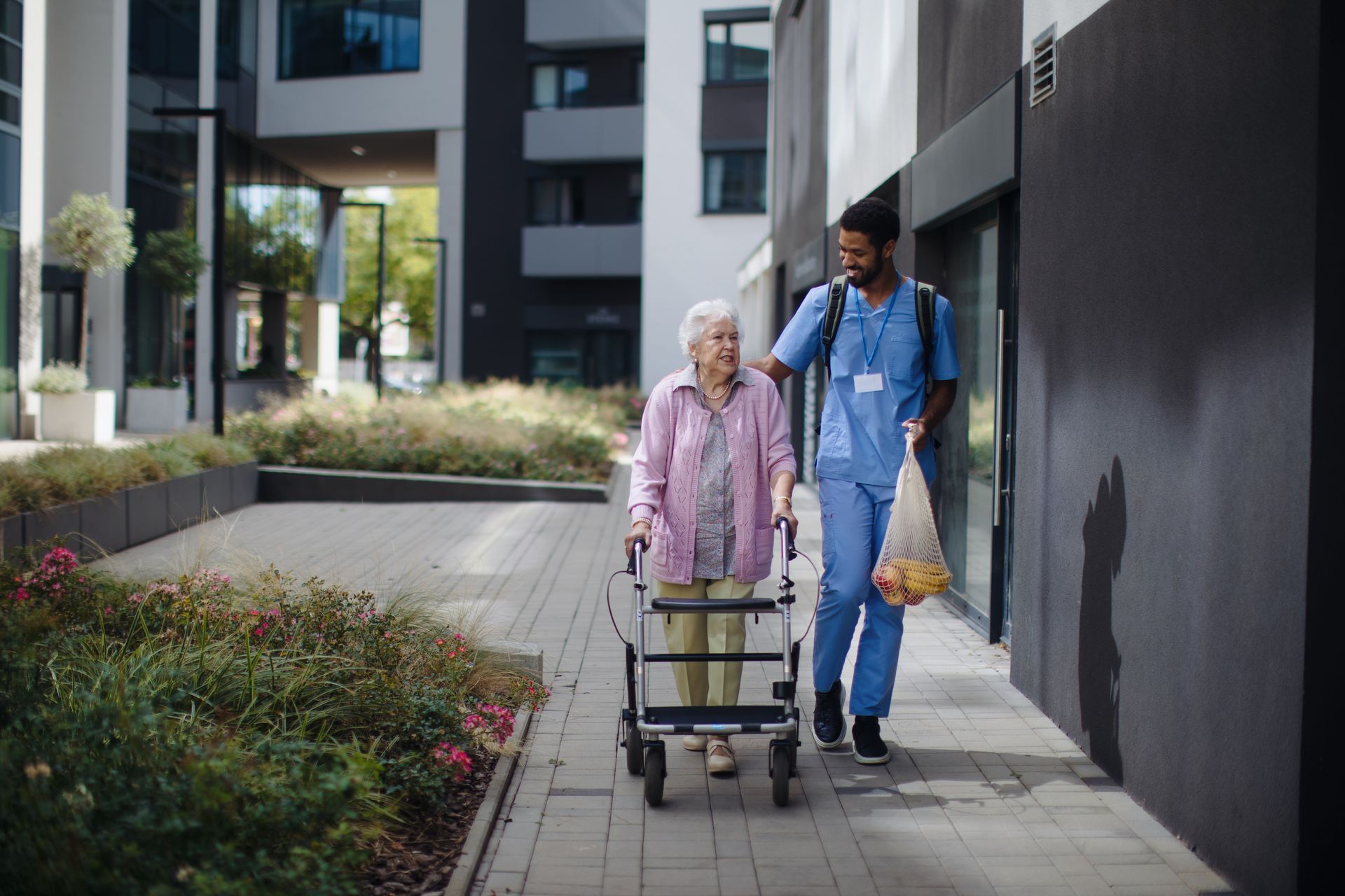 A caregiver assists an elderly woman using a walker along a building walkway.
