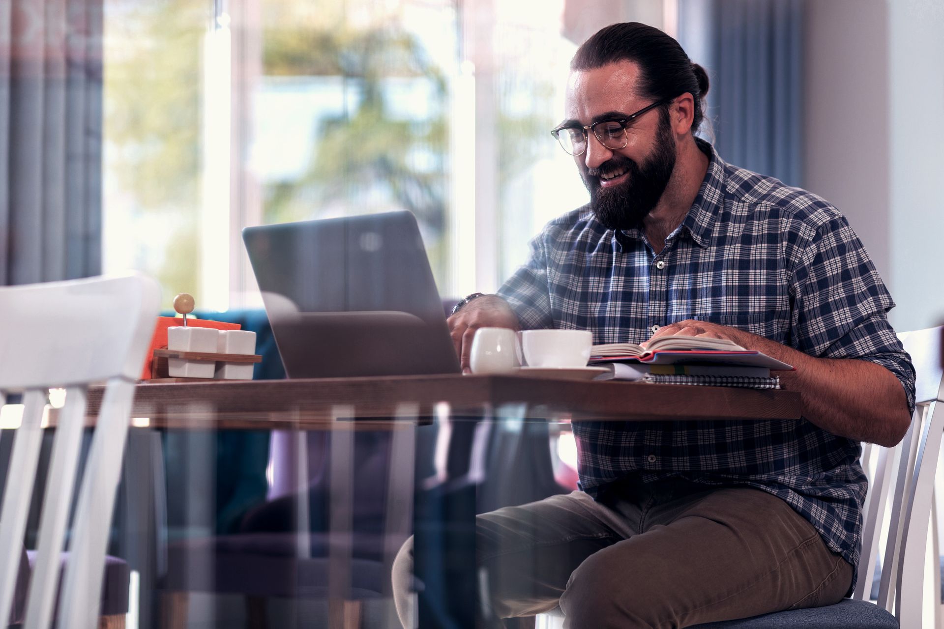 Man with a beard wearing glasses smiles while working on laptop at a cafe table with coffee and food.