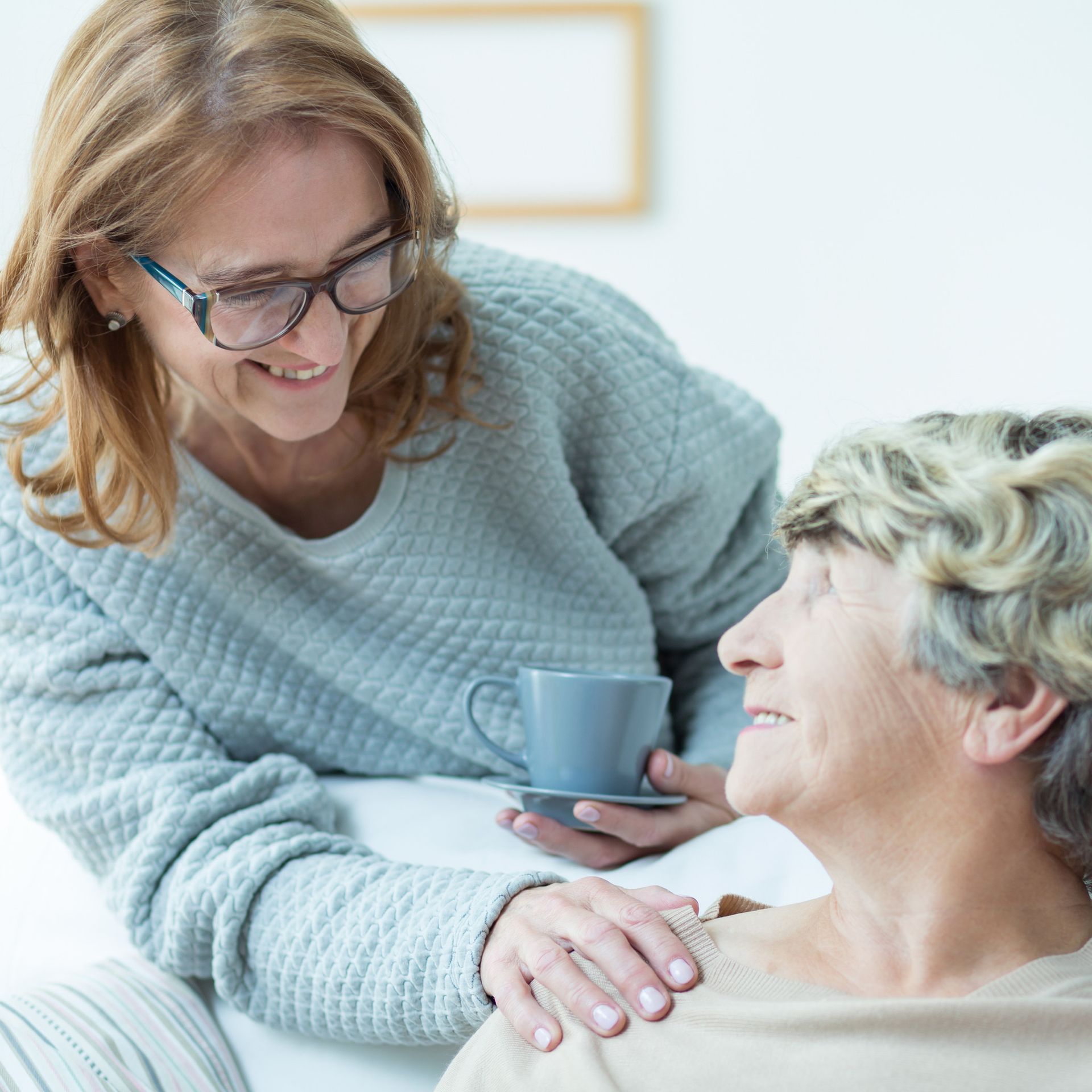Woman in glasses smiles at elderly woman, hand on her shoulder.