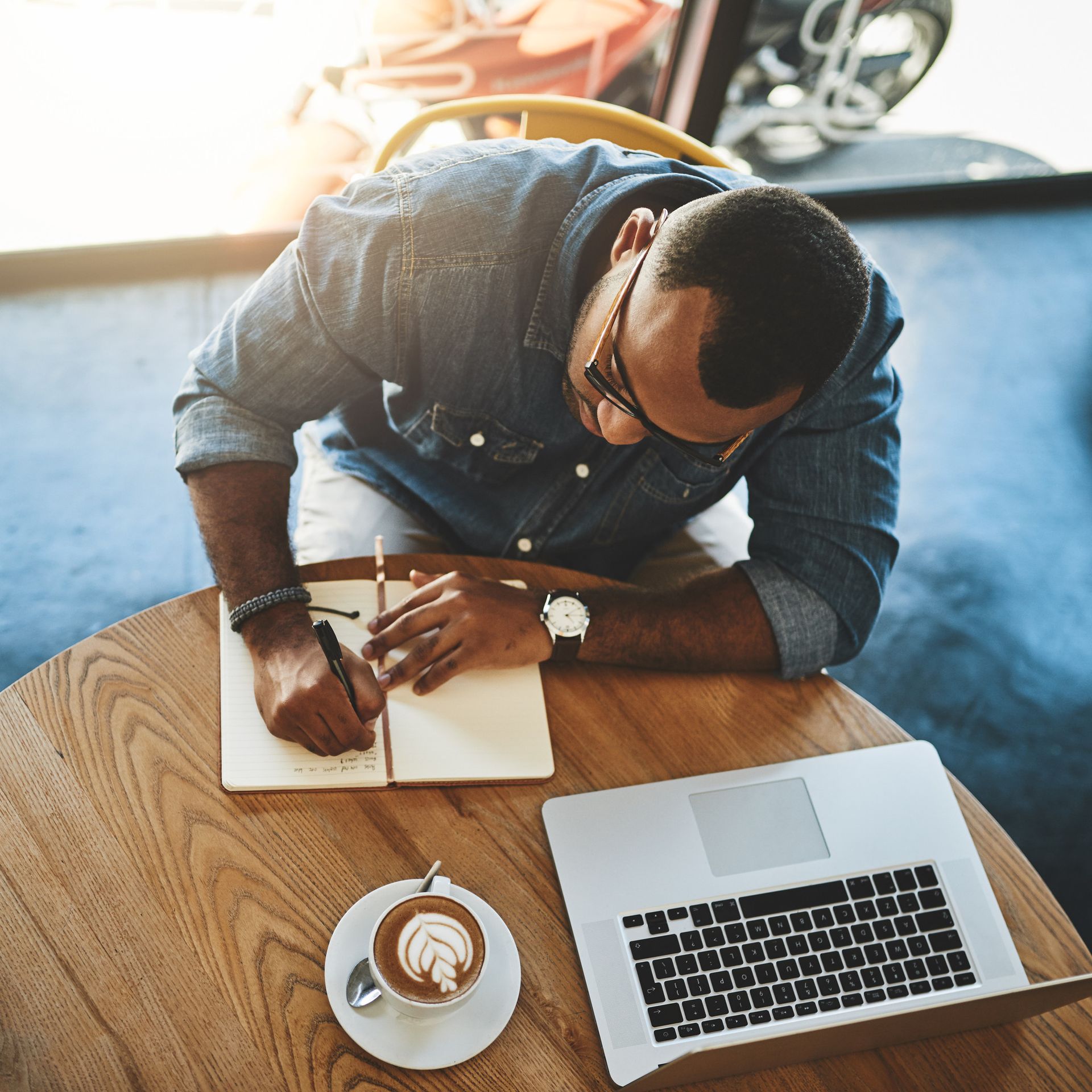 Man in glasses writing in a notebook at a table with a laptop and coffee.
