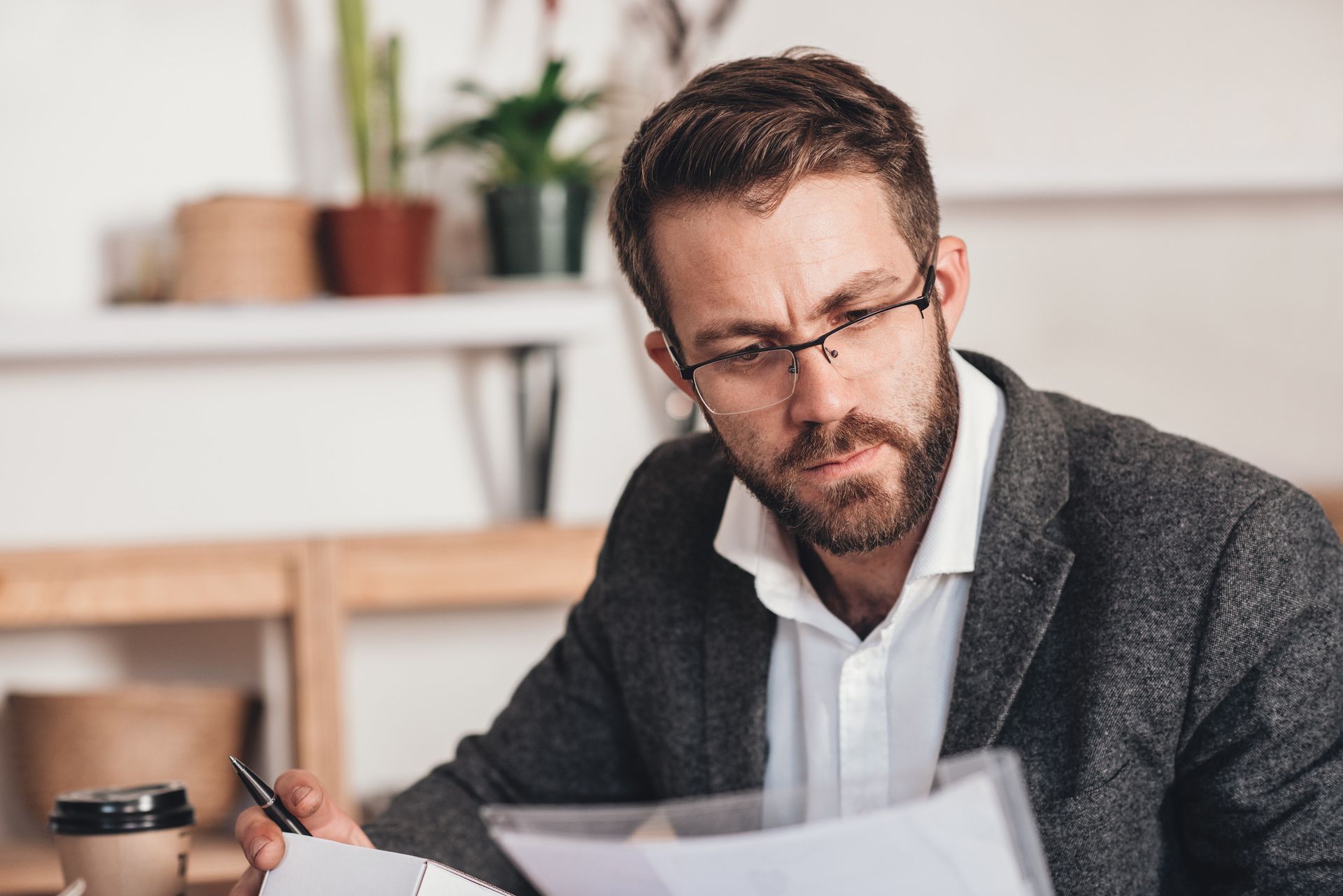 Man with glasses, blazer, and beard intently reviews documents at a desk with a coffee cup.