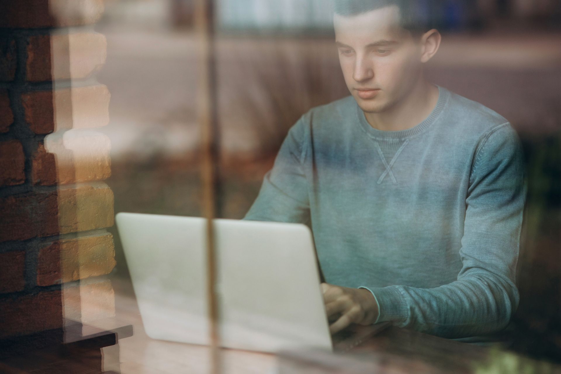 Man using a laptop, seen through a window. He's in a light blue sweater. Brick wall visible.