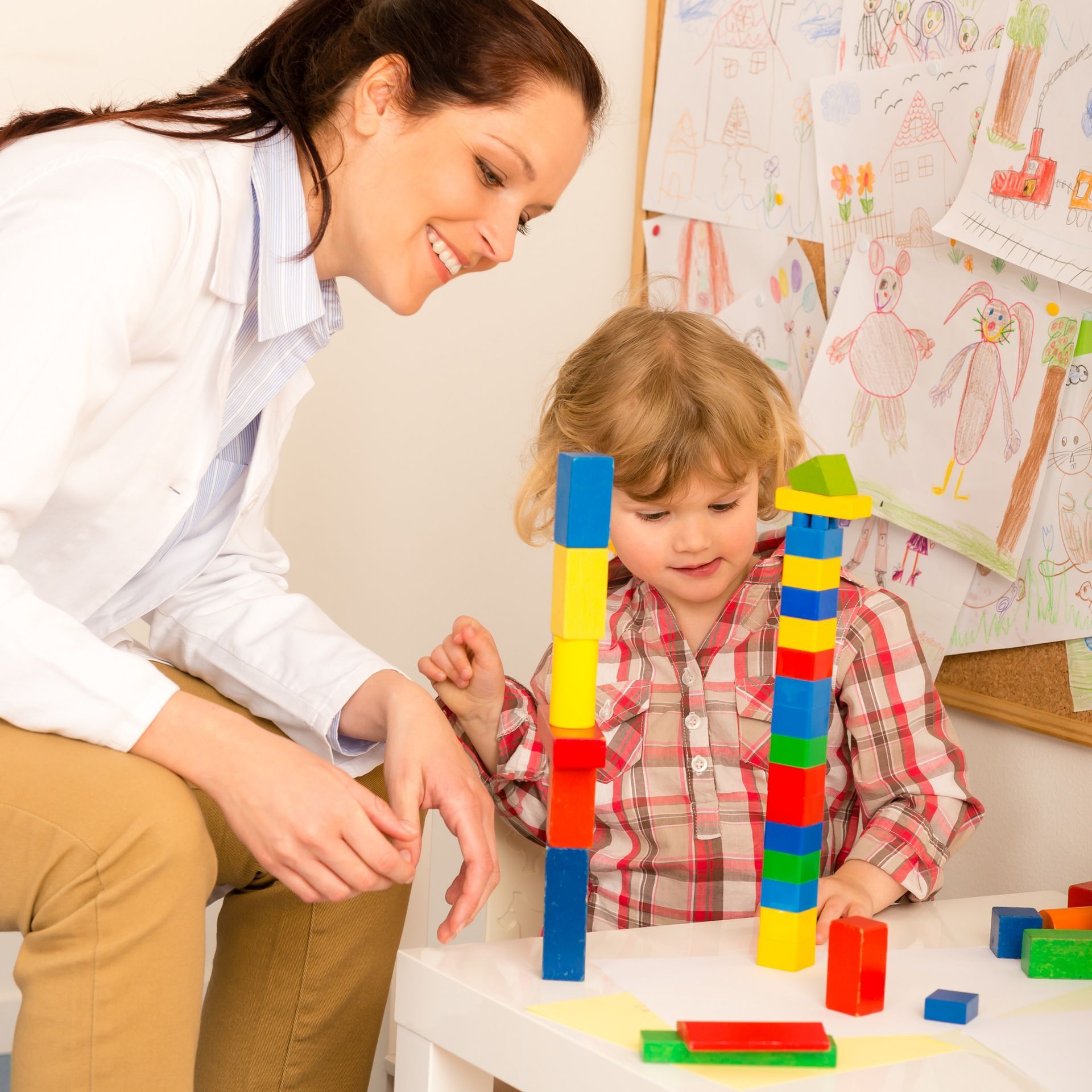 Woman and child building block towers at a table. Woman smiles, observing child's work. Colorful blocks, drawings in background.