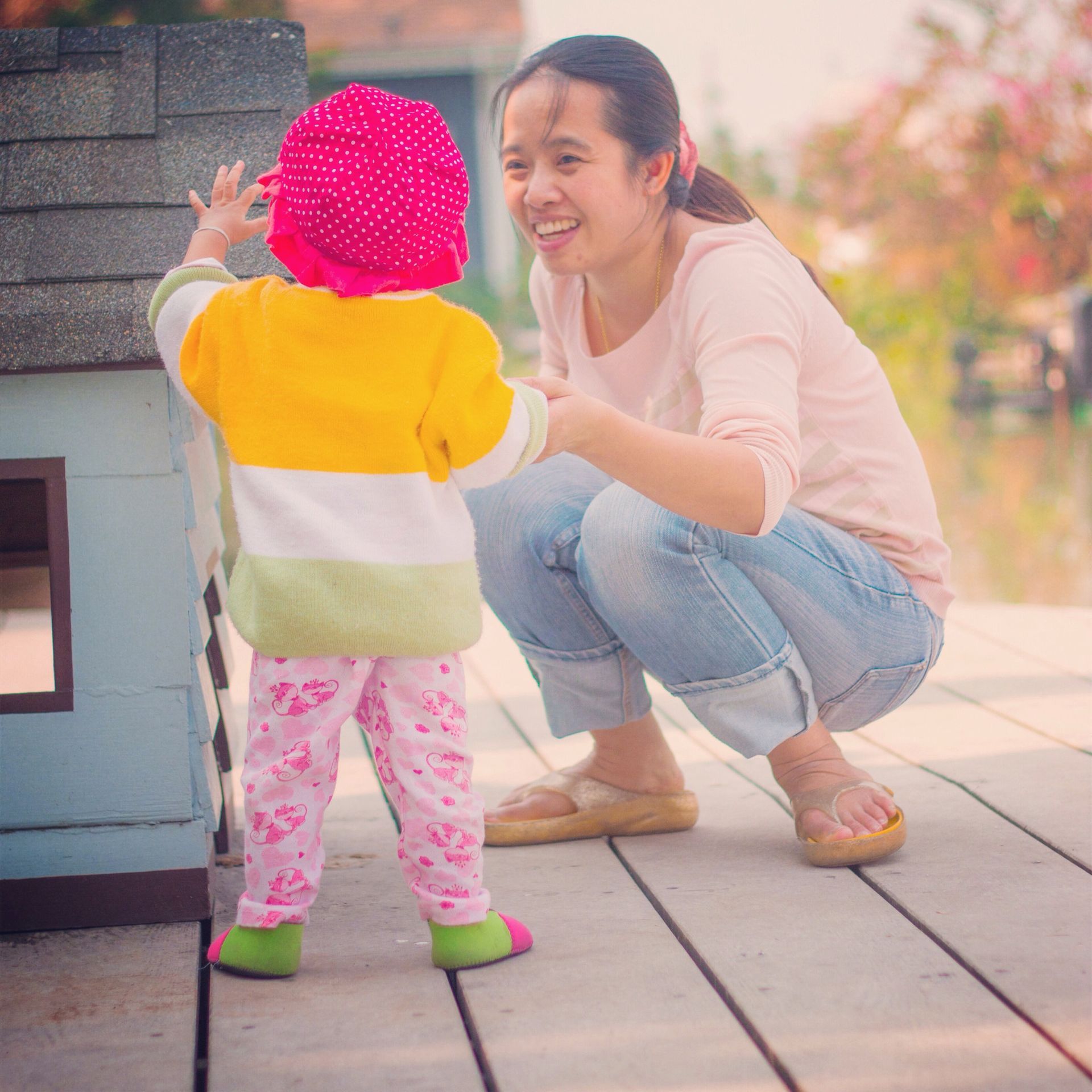 A woman crouches, smiling, reaching toward a toddler in colourful clothes standing near a small playhouse on a wooden deck.