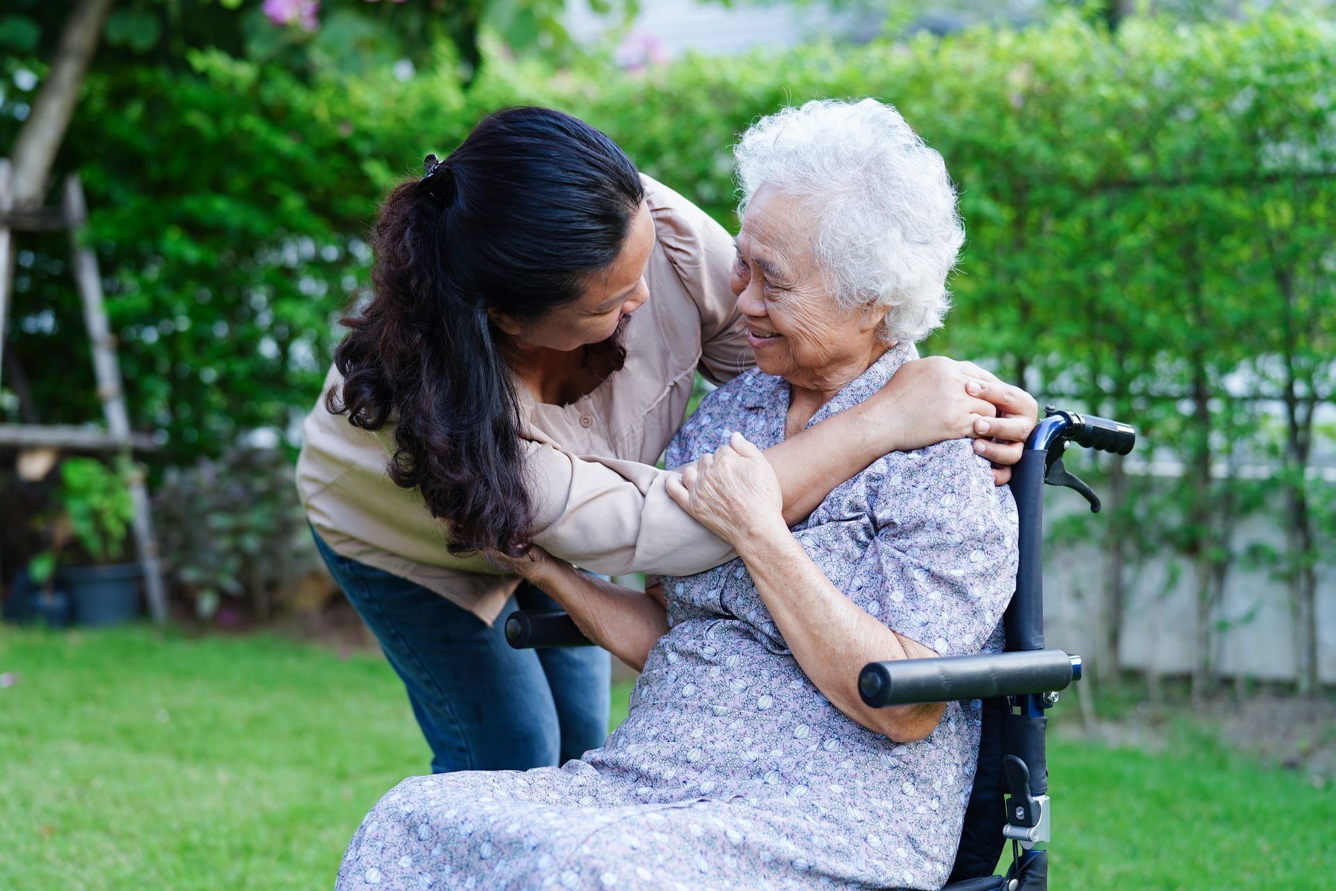 Woman hugging an elderly woman in a wheelchair outdoors. Green grass, trees.