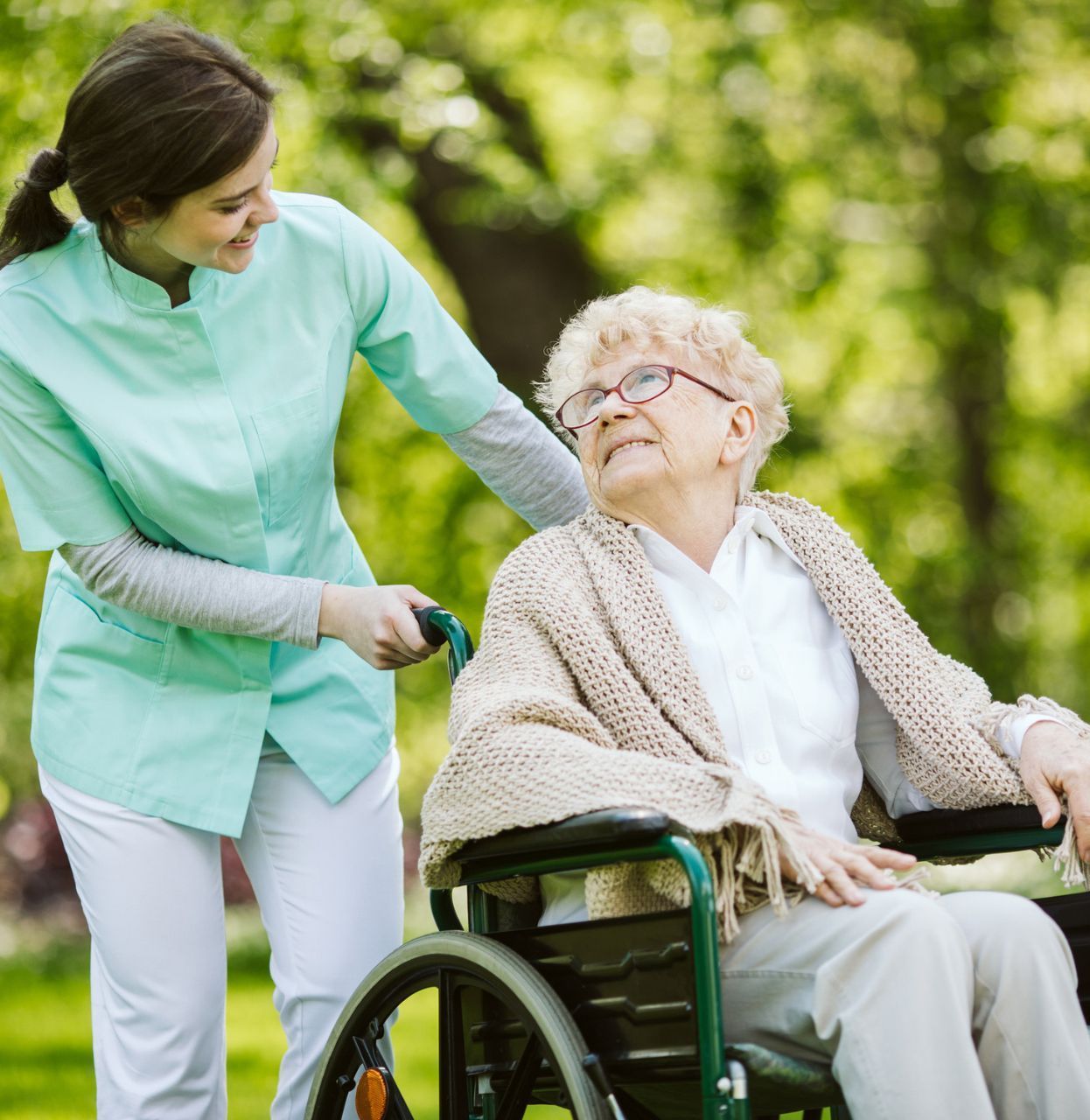 Caregiver pushing an elderly woman in a wheelchair outdoors; both smiling.