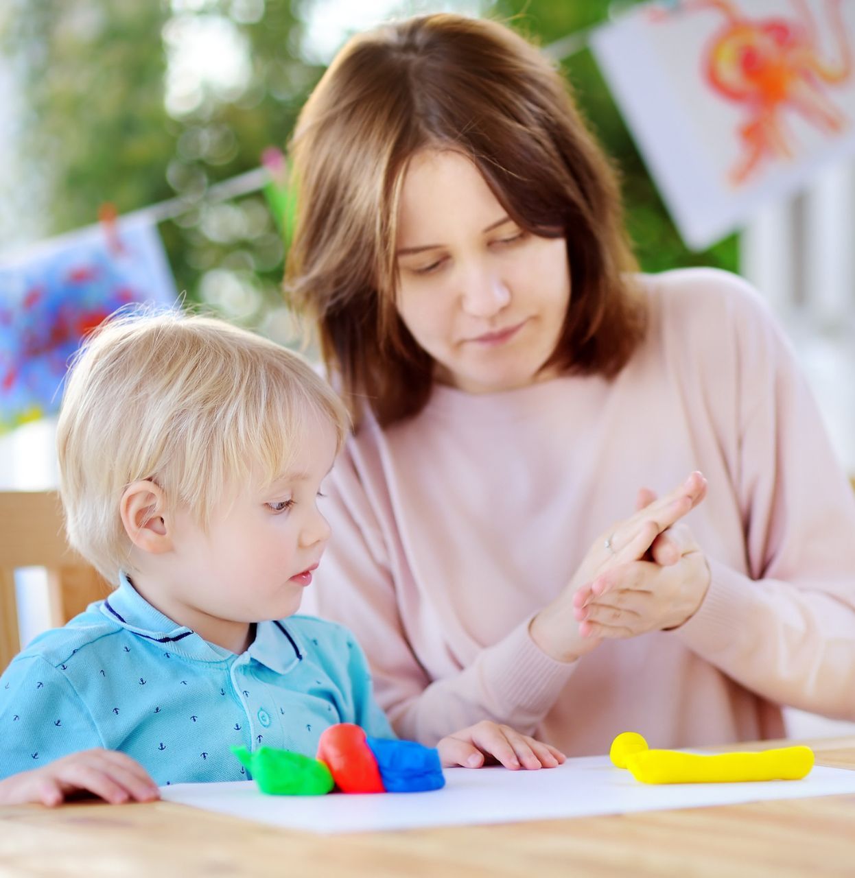 Woman showing a toddler how to play with colorful modeling clay at a table outdoors.