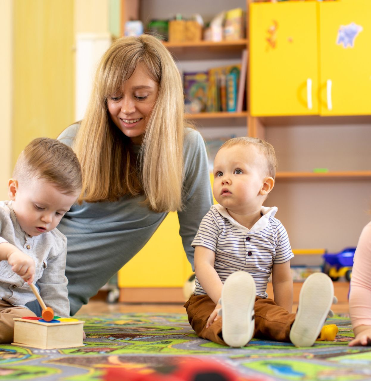 Teacher and three toddlers playing on a colorful rug in a brightly lit room.