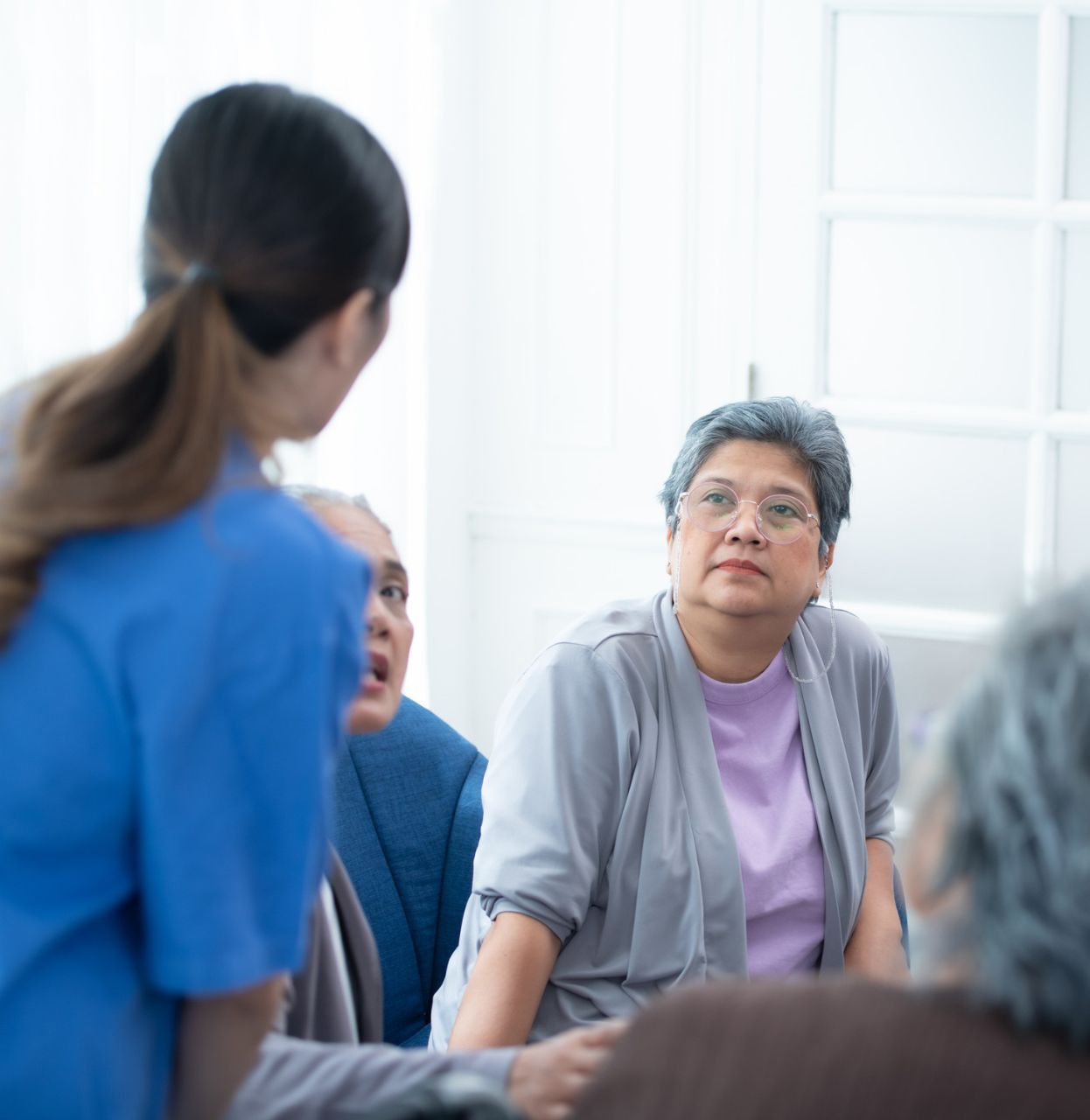 Nurse in blue top speaks to a group of senior women in a bright room.