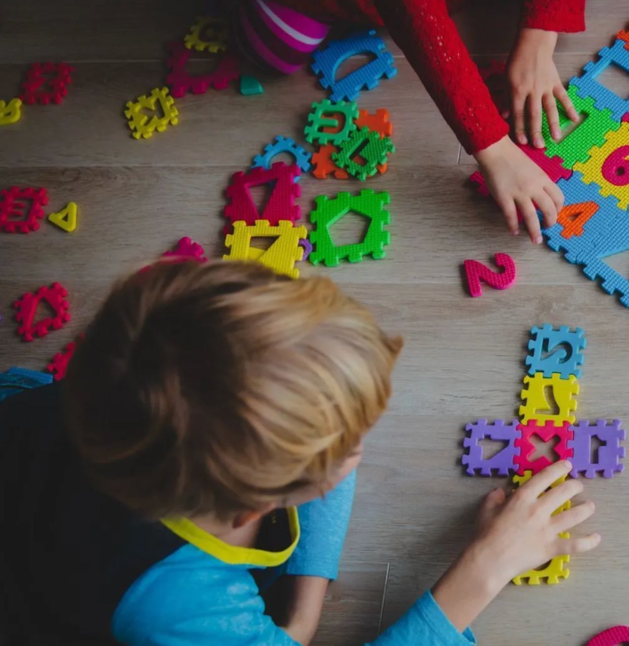 Children playing with colorful foam shape and number puzzle pieces on a wooden floor.