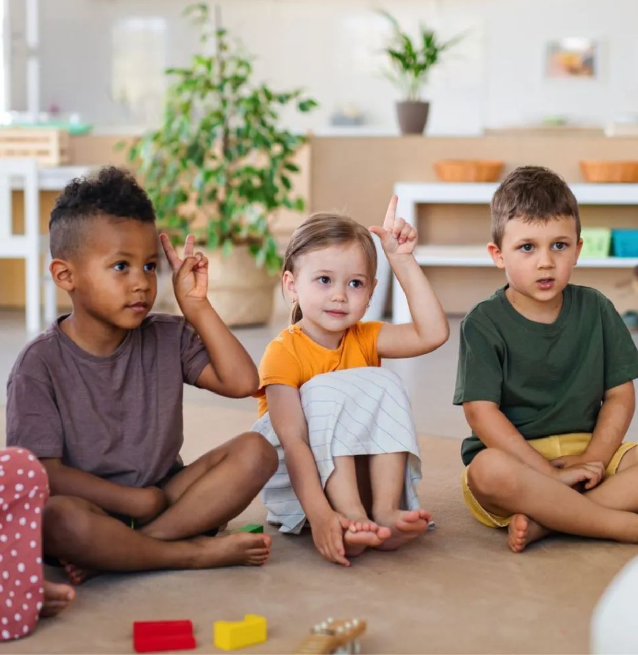 Three children sit on the floor raising their hands. One child looks thoughtful. Bright classroom setting.