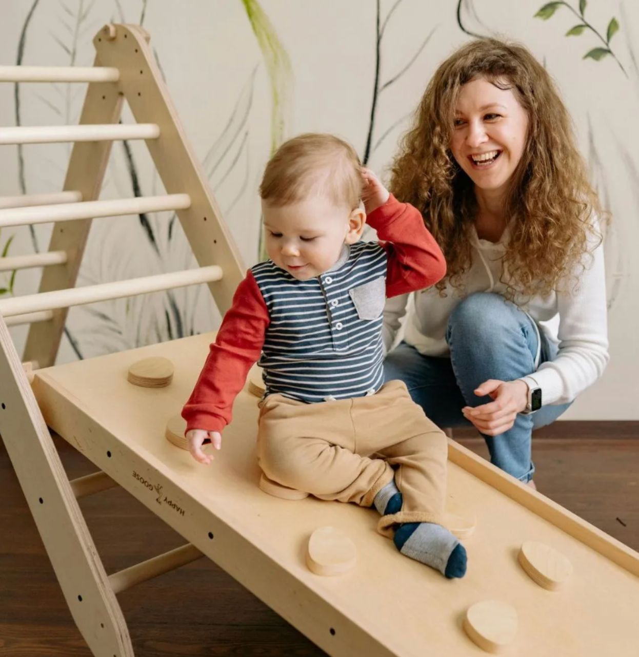 Young child slides down a wooden play structure with assistance from smiling woman.