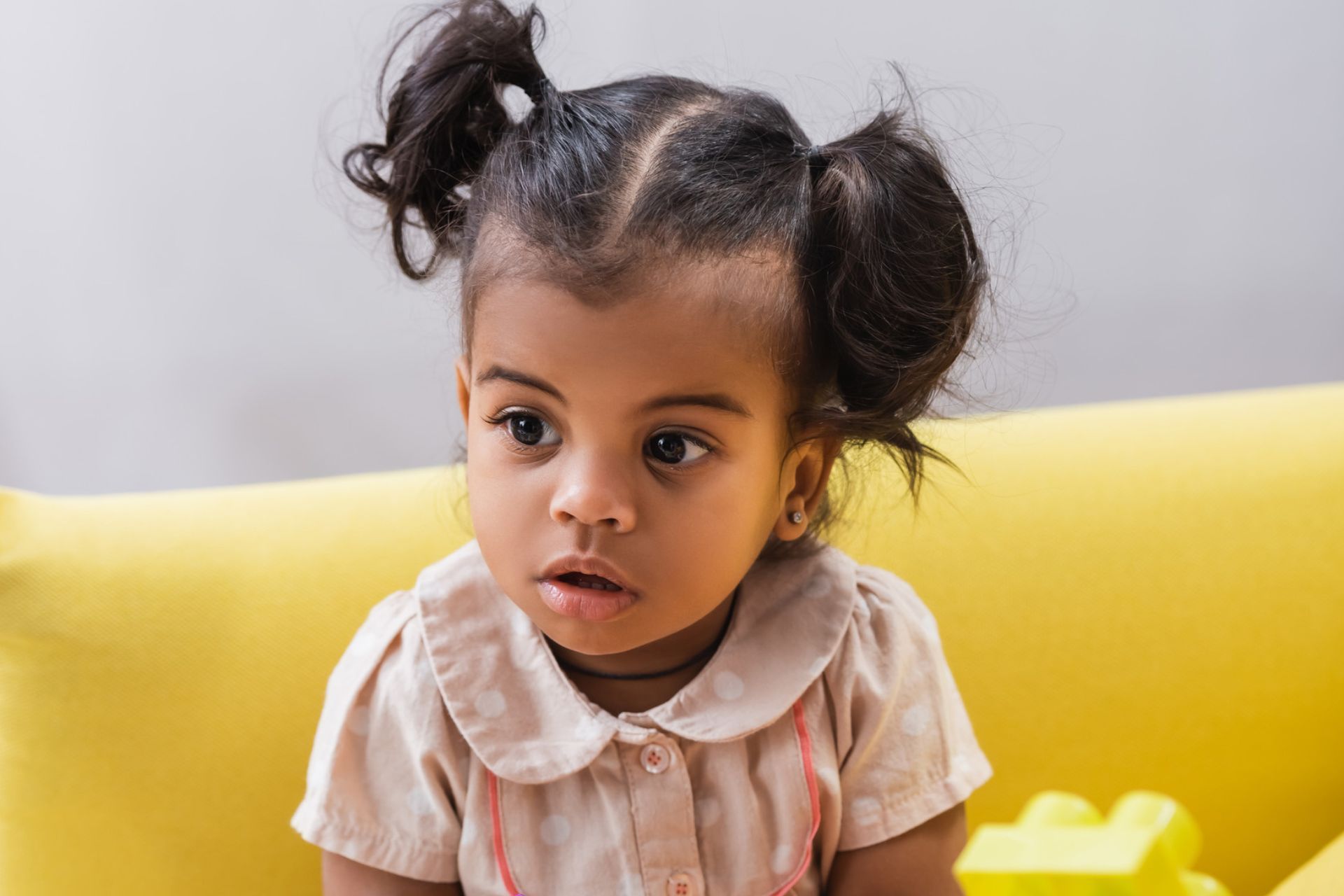 Young Black girl with pigtails, wearing a tan shirt, sits on a yellow couch, looking surprised.