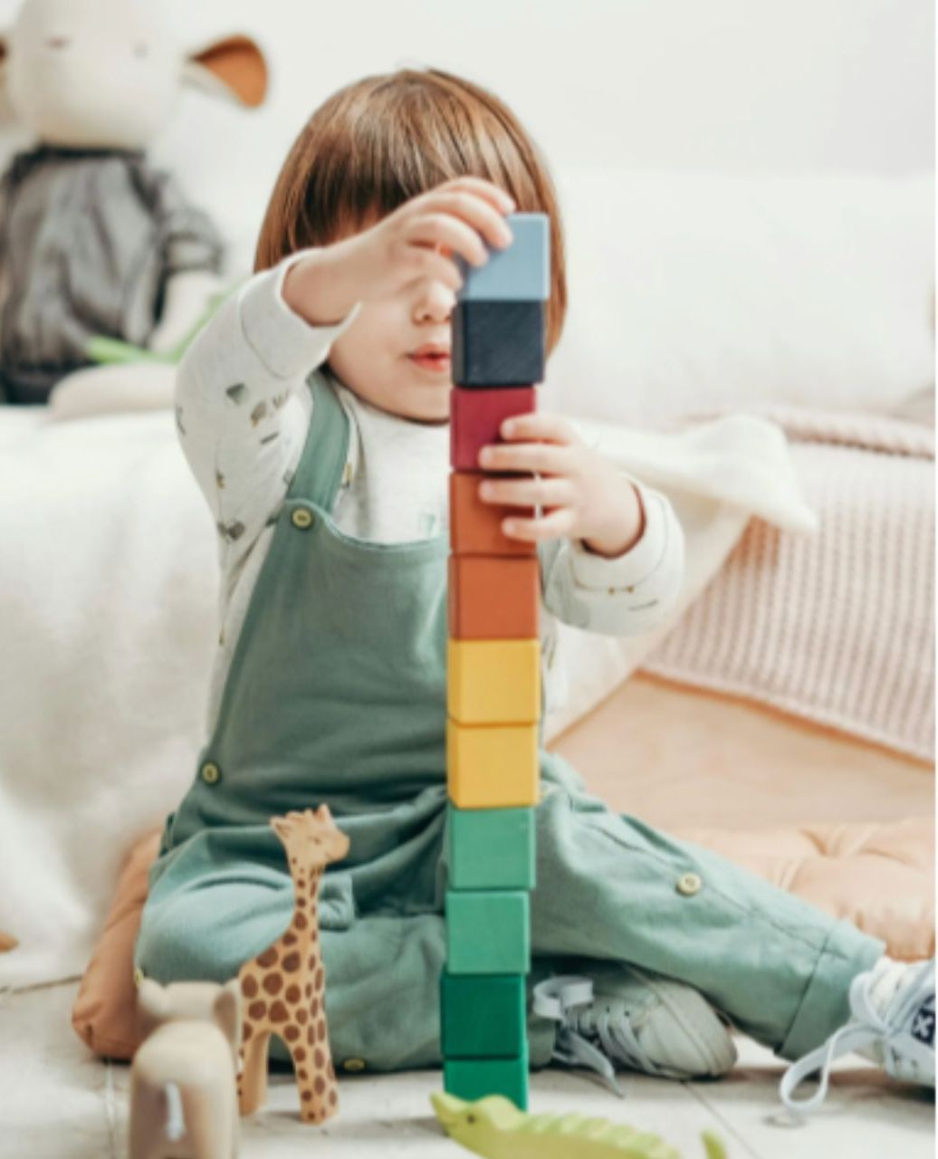 Child in overalls building a colorful tower of blocks on a soft, light-toned floor.