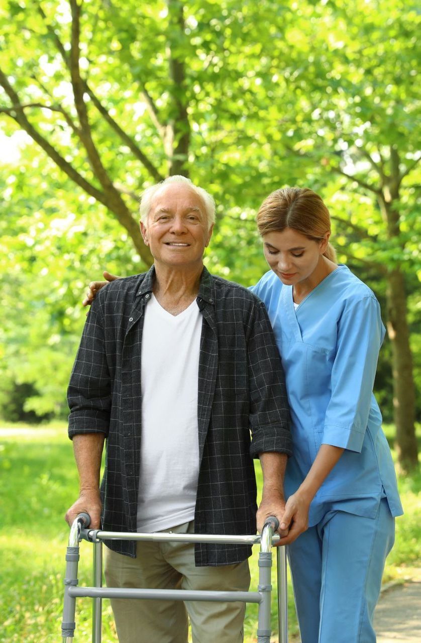 Elderly man using a walker, smiling outdoors with a caregiver in a blue uniform assisting.