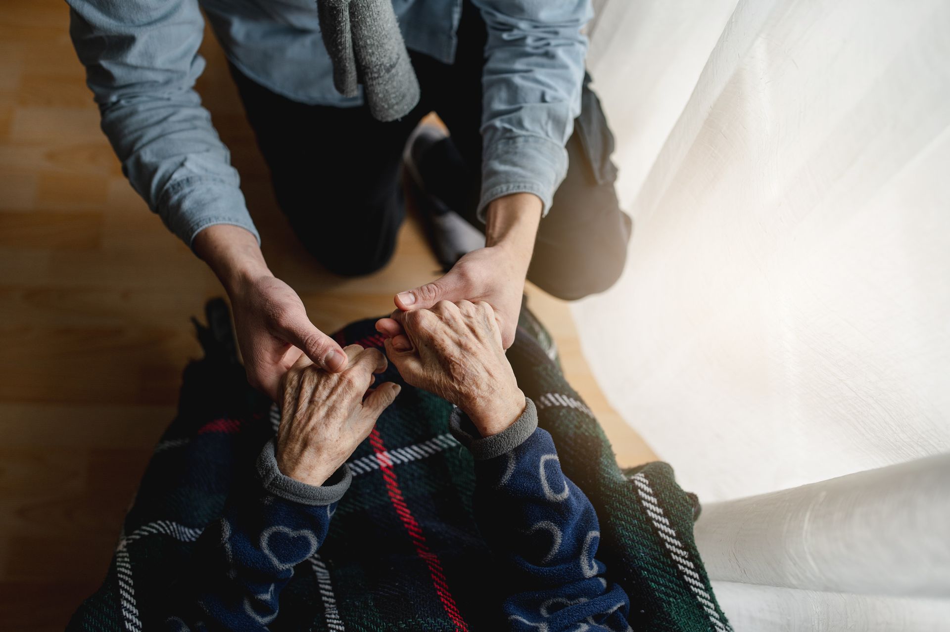 A young person holds an elderly person's hands; indoors, near a window.
