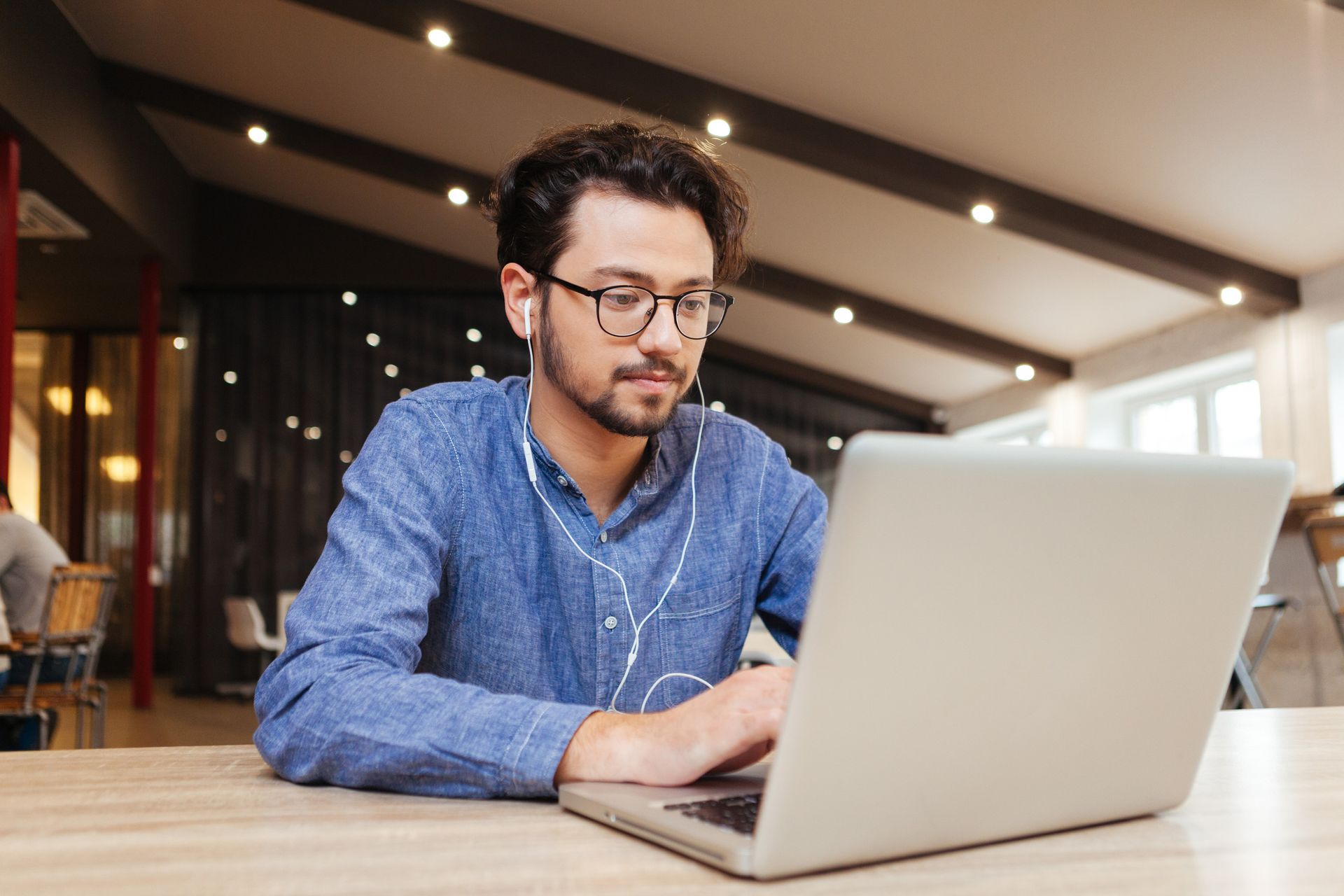 Man with glasses and earbuds, working on a laptop in a modern office setting.