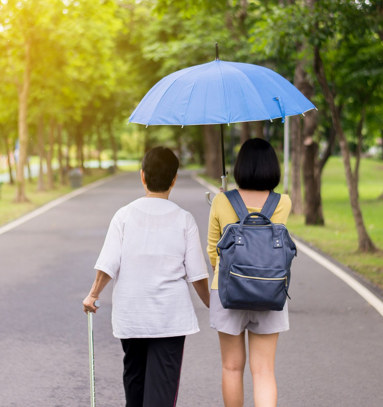 Woman with umbrella walks with older woman using a cane on path in a park.