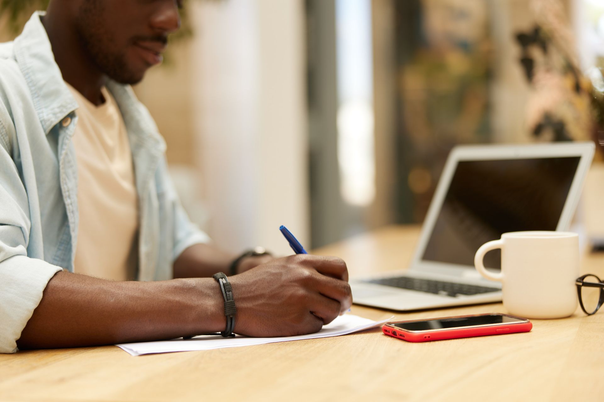 Black man writing notes at a table with a laptop, mug, and phone, indoors.