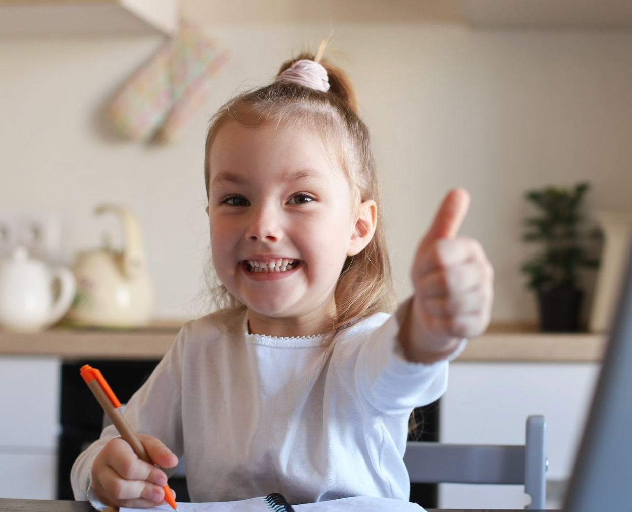 Smiling young girl giving thumbs up while writing at a table in a kitchen.