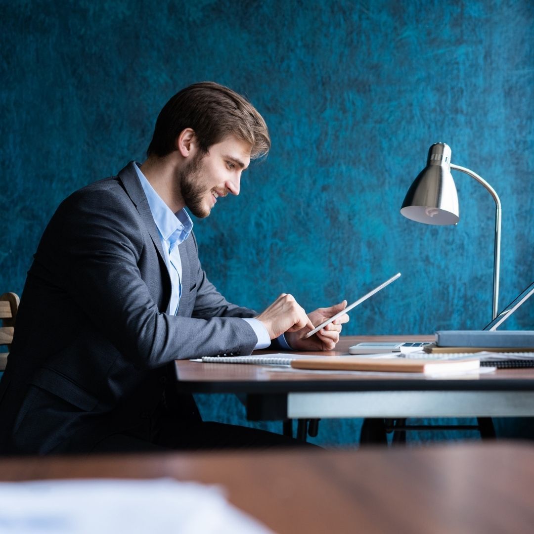 Man in suit smiles, using a tablet at a desk. Blue background, lamp lit.