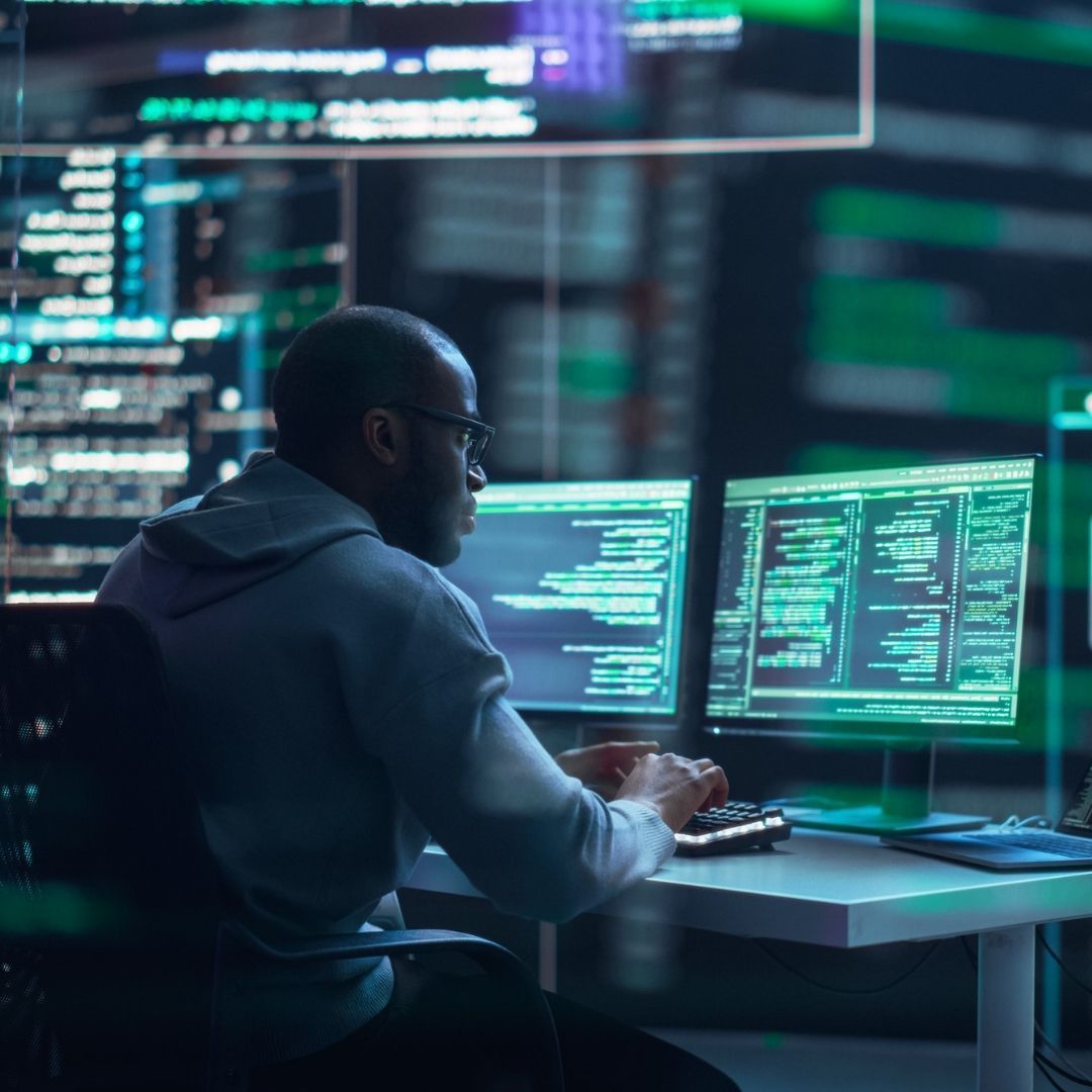 Man in glasses working on computer code, lit by screens in a dark setting.