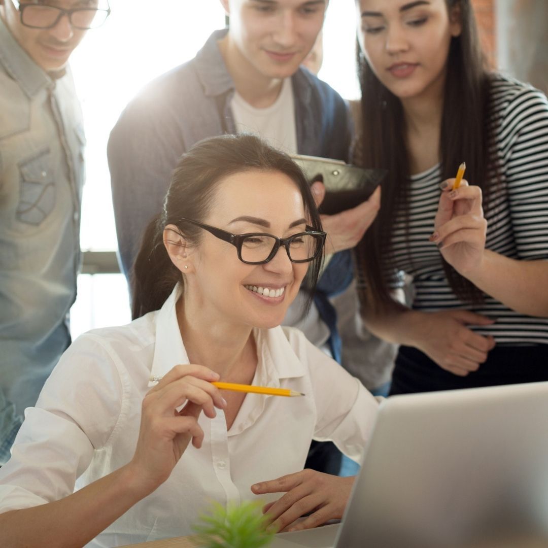 Woman in glasses smiles at laptop, surrounded by colleagues in a bright office setting.