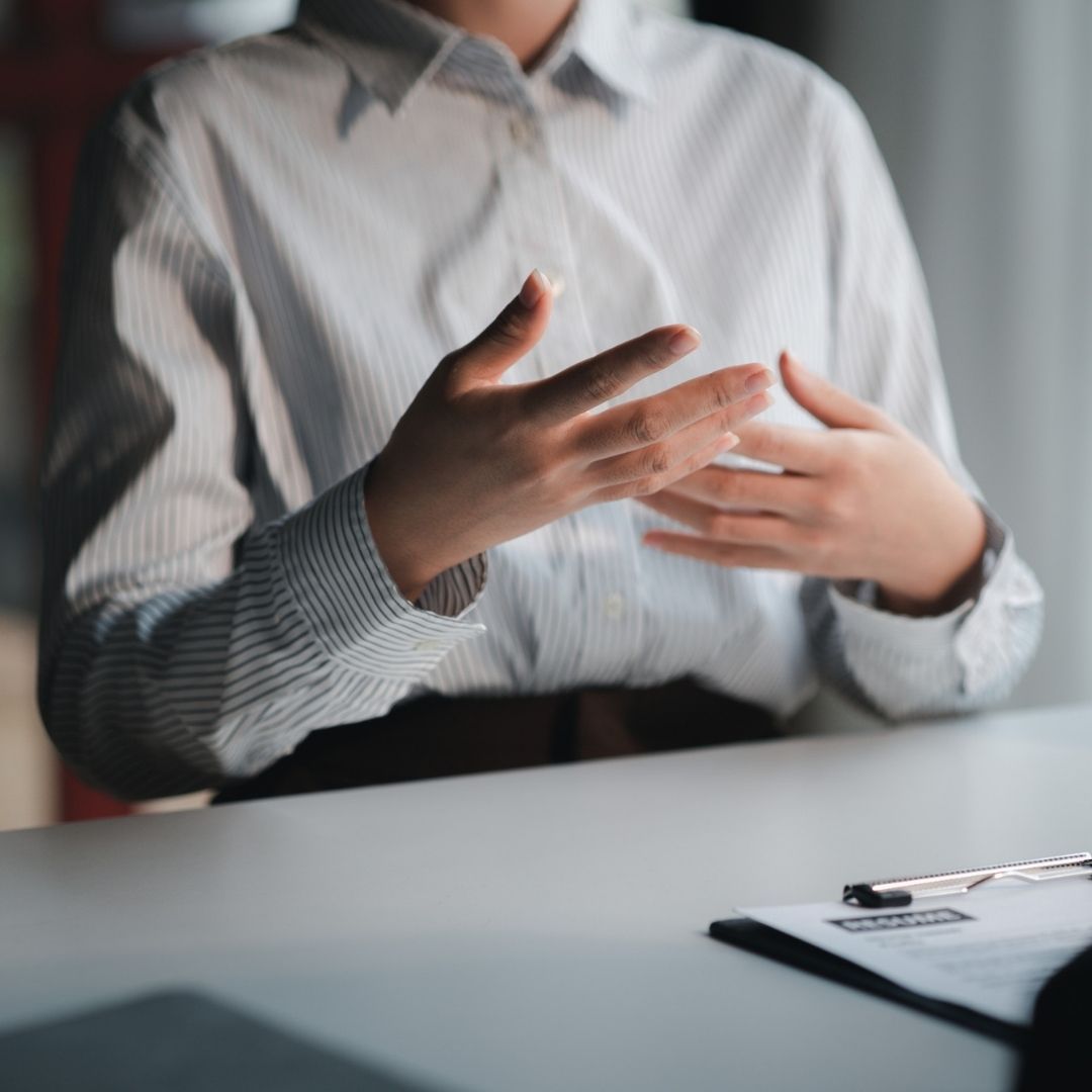 Person in white shirt gestures with hands, sitting at a desk with a clipboard.