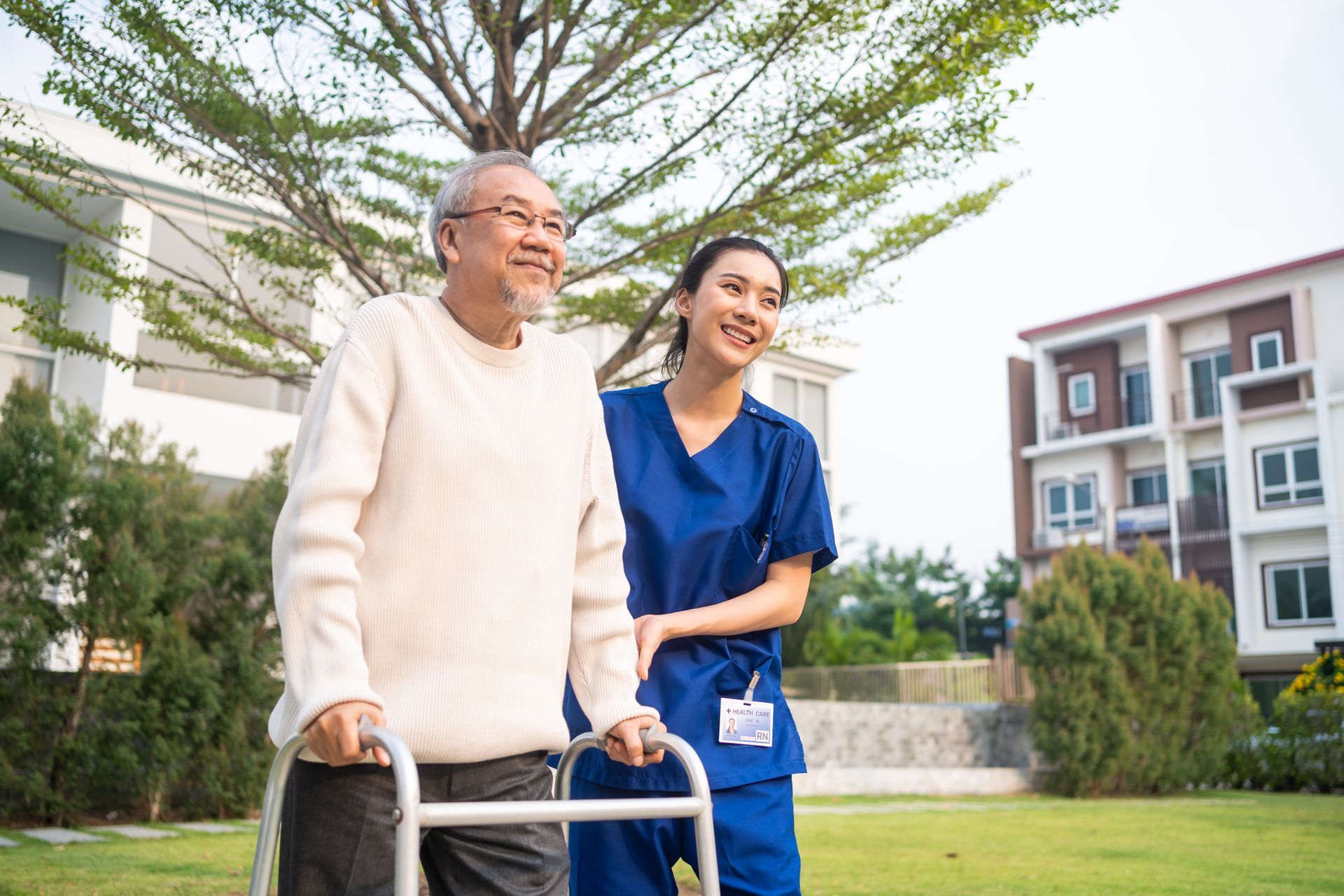 An older Asian man uses a walker, smiling while walking with a caregiver outdoors.