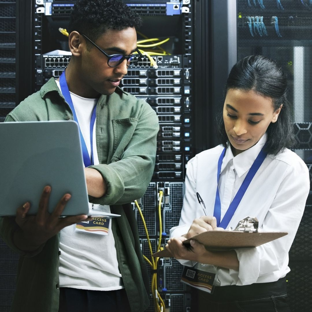 Two IT technicians working in a server room, one on a laptop, the other taking notes.