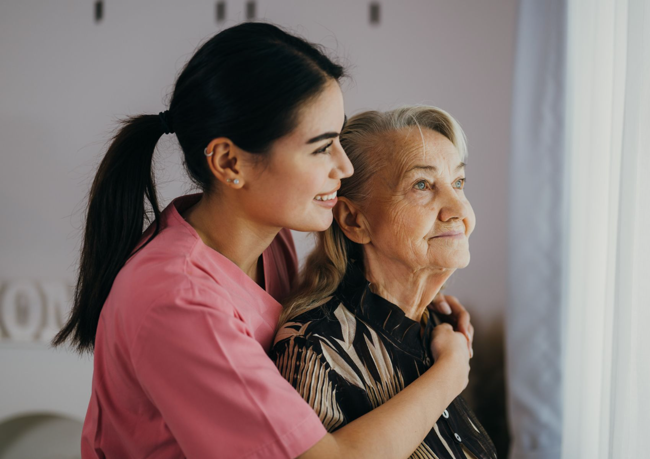 Young woman in pink shirt hugs older woman looking out a window.