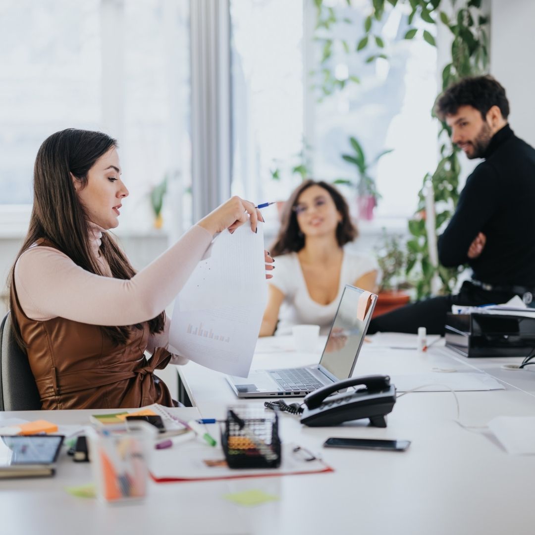 Woman in brown top reviews papers, facing two colleagues at an office desk.