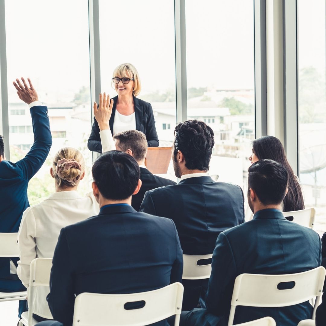 Businesswoman presenting to a group, some with hands raised in a bright room with a city view.